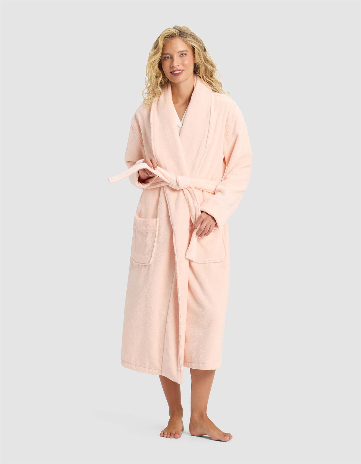 A smiling woman with blonde hair stands barefoot, facing forward in the Cozy Earth Luxe Bath Robe—a long, light pink robe with front pockets and a tie belt—set against a plain light gray background.