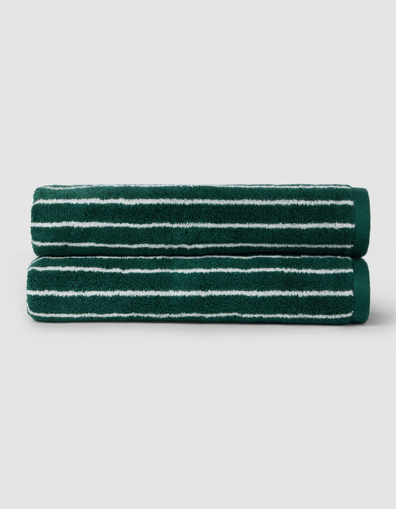 Two Luxe Bath Towels by Cozy Earth, dark green with white horizontal stripes, are neatly rolled and stacked on a light gray background.