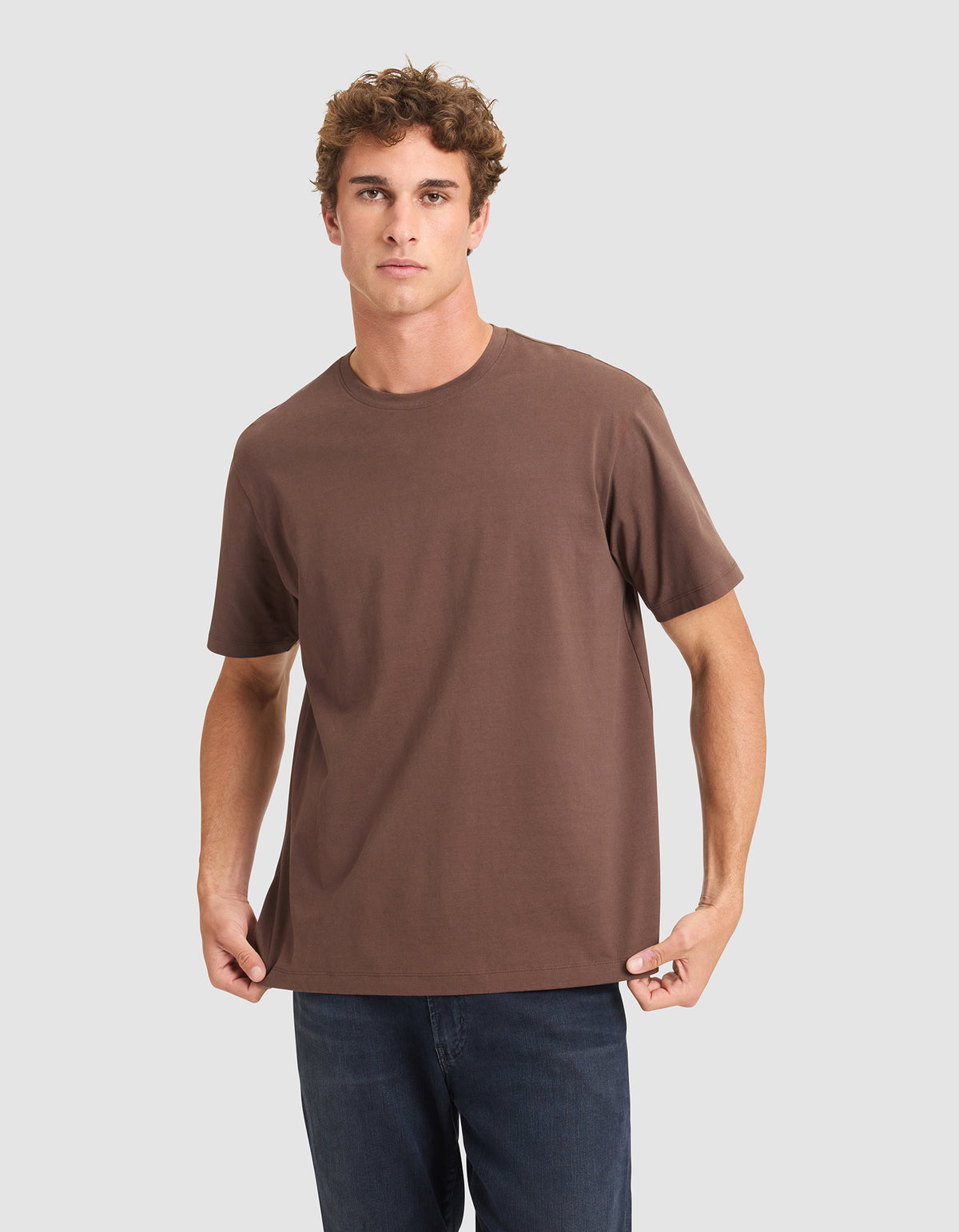 A young man with curly brown hair wears the Cozy Earth Men's All Day Tee, a loose-fitting brown short-sleeve shirt, paired with dark blue jeans against a light gray background. 