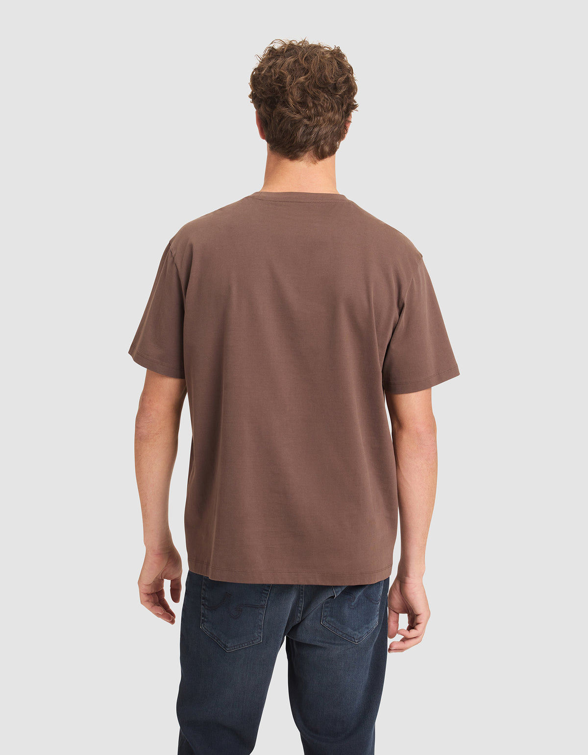 A man with short curly hair, seen from behind, wears a Cozy Earth Men's All Day Tee in brown and dark blue jeans against a plain light background. 
