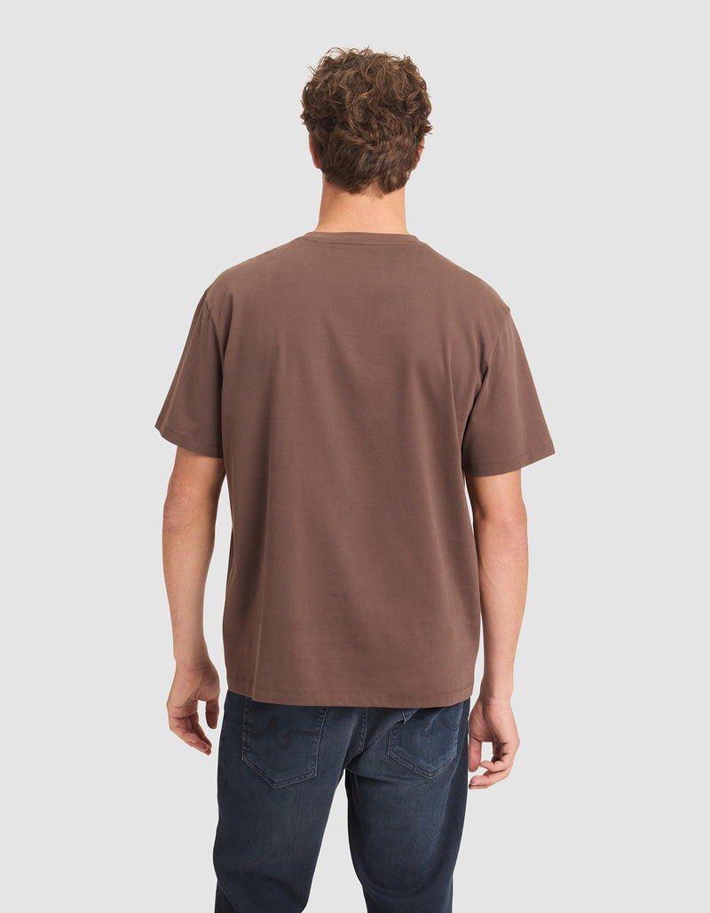 A man with short curly hair, seen from behind, wears a Cozy Earth Men's All Day Tee in brown and dark blue jeans against a plain light background.