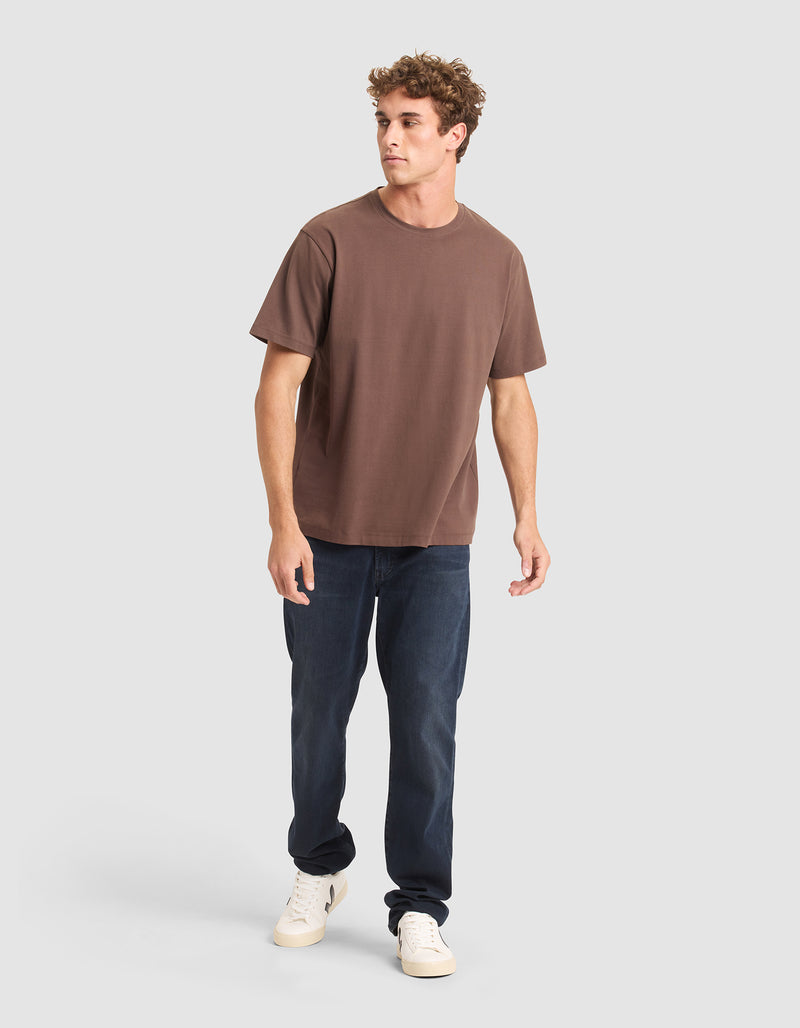A young man stands on a light gray background wearing the Cozy Earth Men's All Day Tee in brown, paired with dark blue jeans and white sneakers. He looks slightly to the side, one leg forward as if walking.