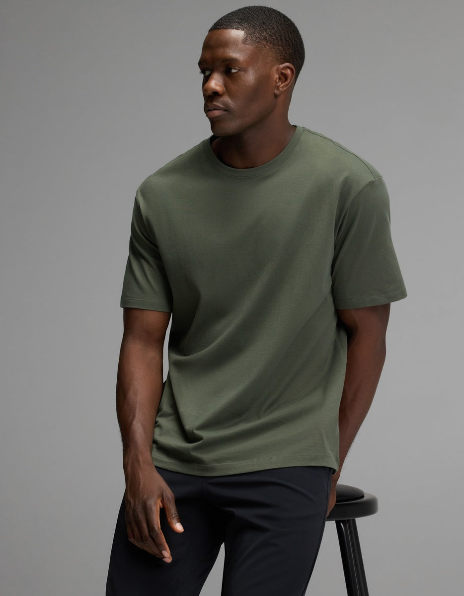 A man wearing the Cozy Earth Men’s All Day Tee in olive and black pants sits on a black stool against a plain gray background, looking to his left with a neutral expression.