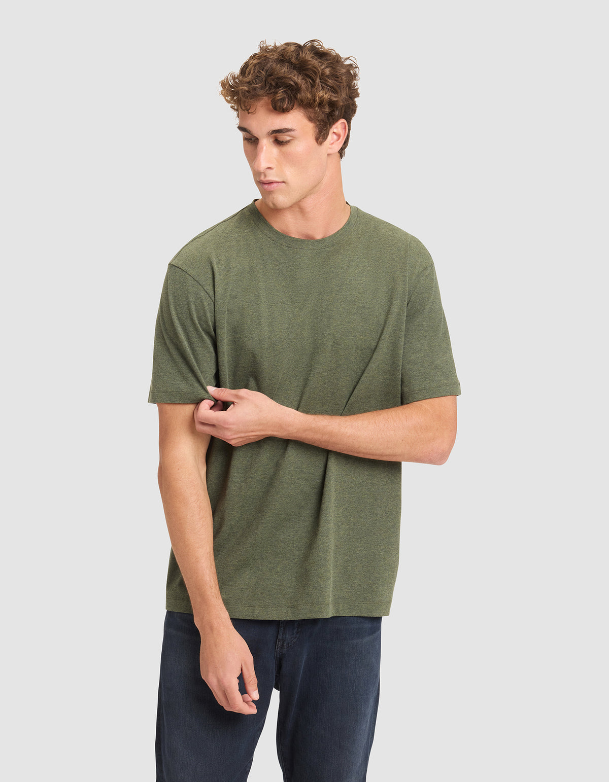 A young man with curly hair wears the Cozy Earth Men's All Day Tee in olive green and dark jeans, standing against a plain light gray background while looking down and gently pulling his sleeve. 