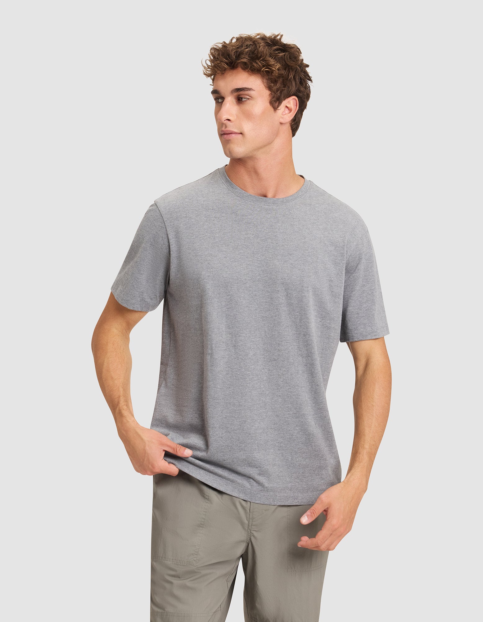 A young man with curly hair wears the Cozy Earth Men's All Day Tee in grey and light khaki pants, standing against a white background with one hand in his pocket and the other adjusting his shirt. |Color:Stone Heather