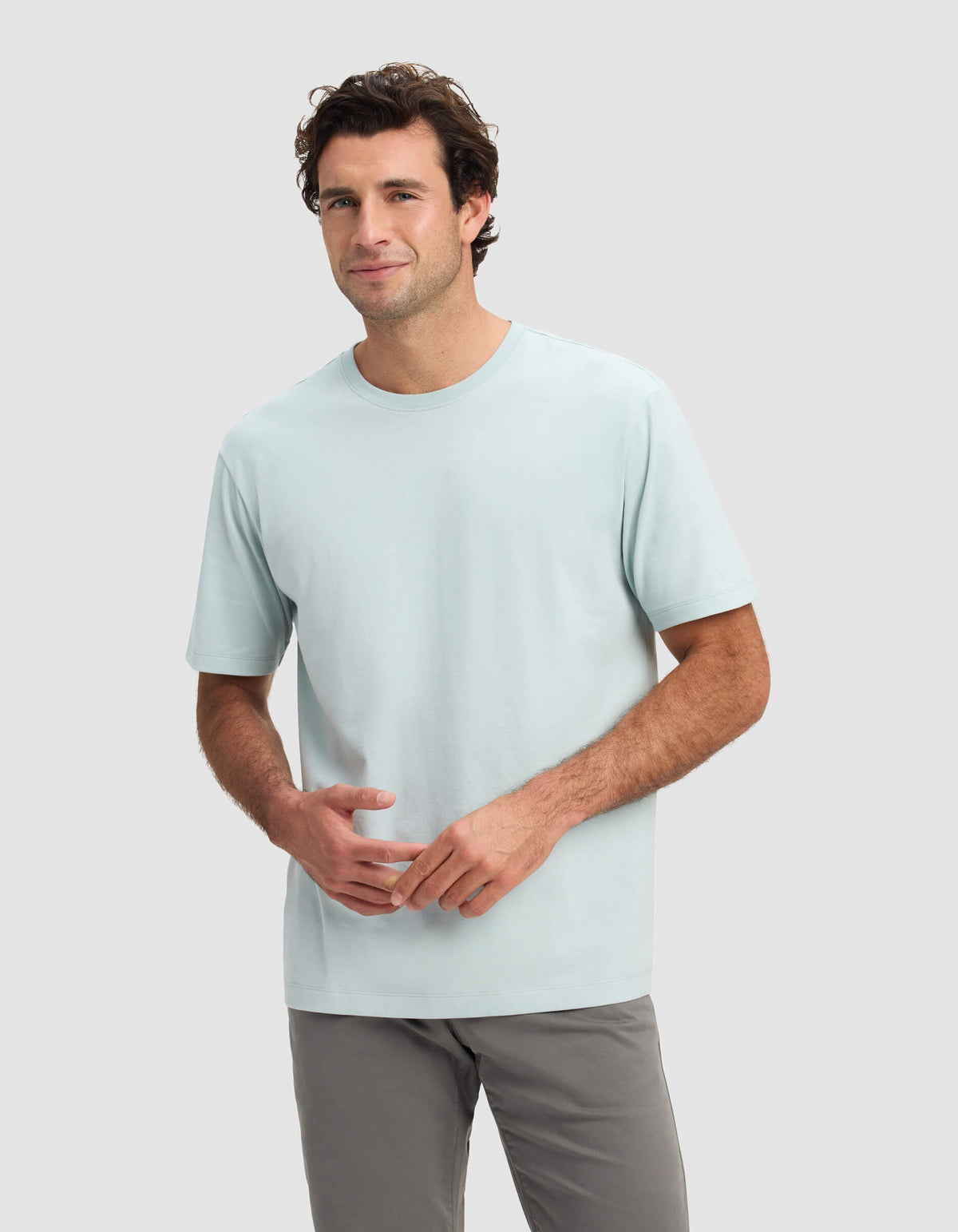 A man with short brown hair is wearing the Cozy Earth Men's All Day Tee in light blue and gray pants, standing against a plain light gray background and smiling slightly.
