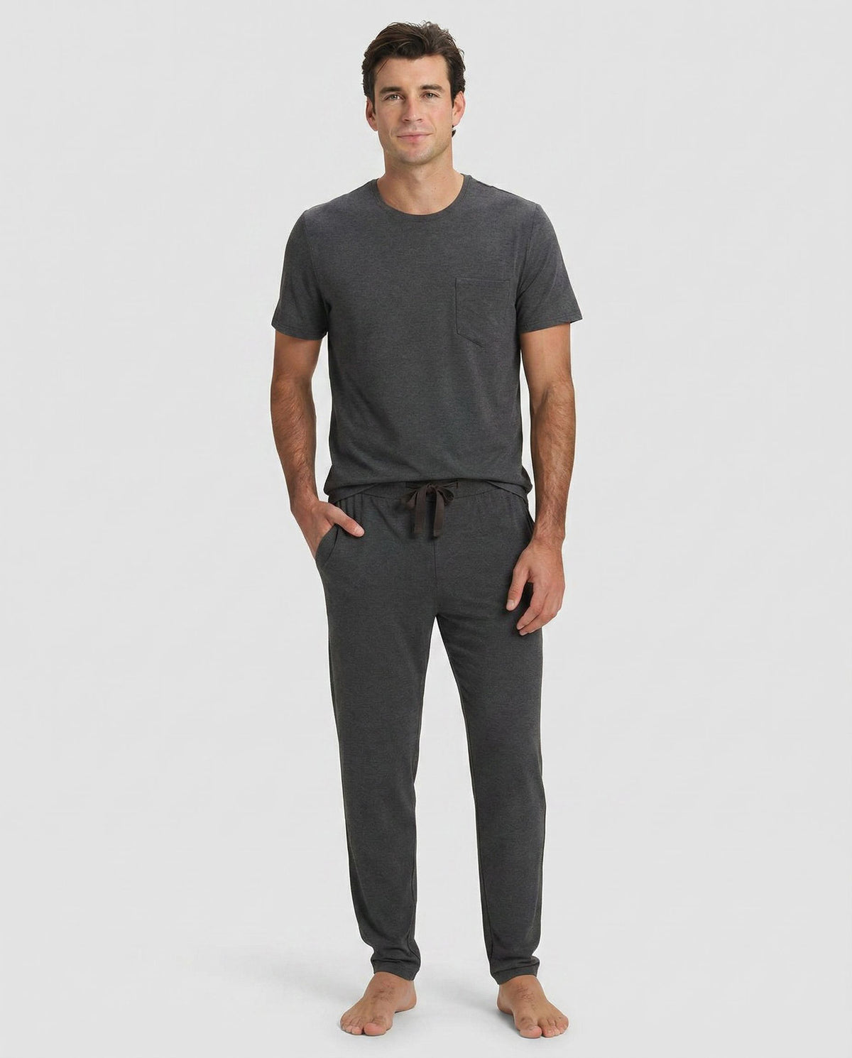 A man stands barefoot against a plain light background, wearing Cozy Earth’s Men's Bamboo Stretch-Knit Lounge Tee & Pant Pajama Set in Charcoal.|Color:Charcoal