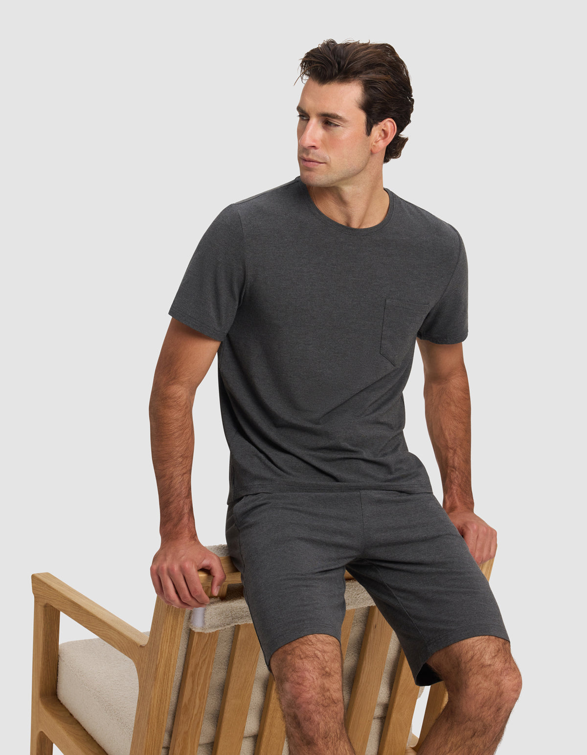 A man wears the Cozy Earth Men's Bamboo Stretch-Knit Lounge Tee in dark gray, paired with matching shorts, while sitting sideways on a light wooden chair with beige cushions against a plain light background.