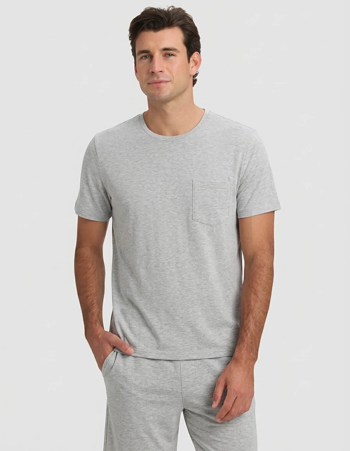 A man models the Cozy Earth Men's Bamboo Stretch-Knit Short Sleeve Pajama Set—light gray with a chest pocket and matching pants—standing with one hand in his pocket against a plain light background.
