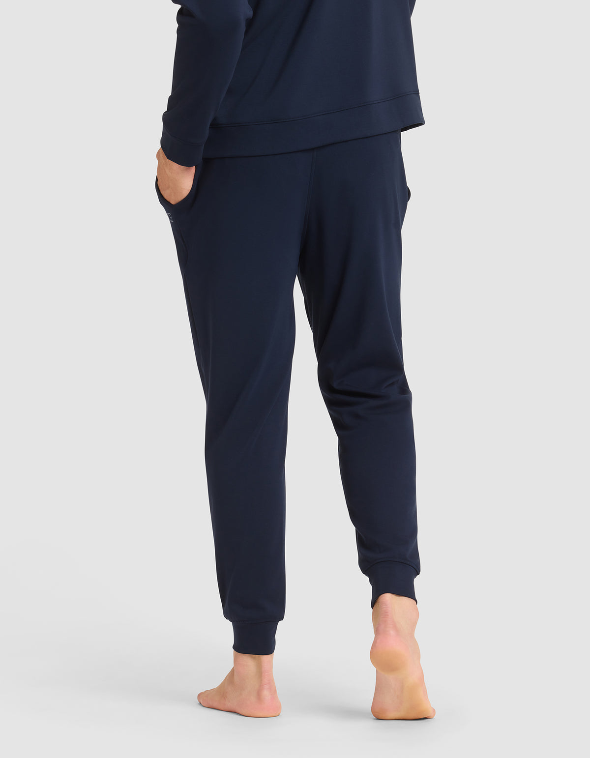 A person stands barefoot on a white surface, shown from the back wearing Cozy Earth's Men's Brushed Bamboo Jogger Pant in navy blue with a matching long-sleeve top. One hand is in a pocket and the left heel is slightly lifted.