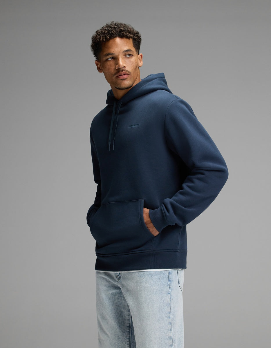 A man wearing the Cozy Earth Men’s Cityscape Hoodie in Eclipse stands against a plain gray background with his hands in the front pocket, looking to the side, and styled with light blue jeans.