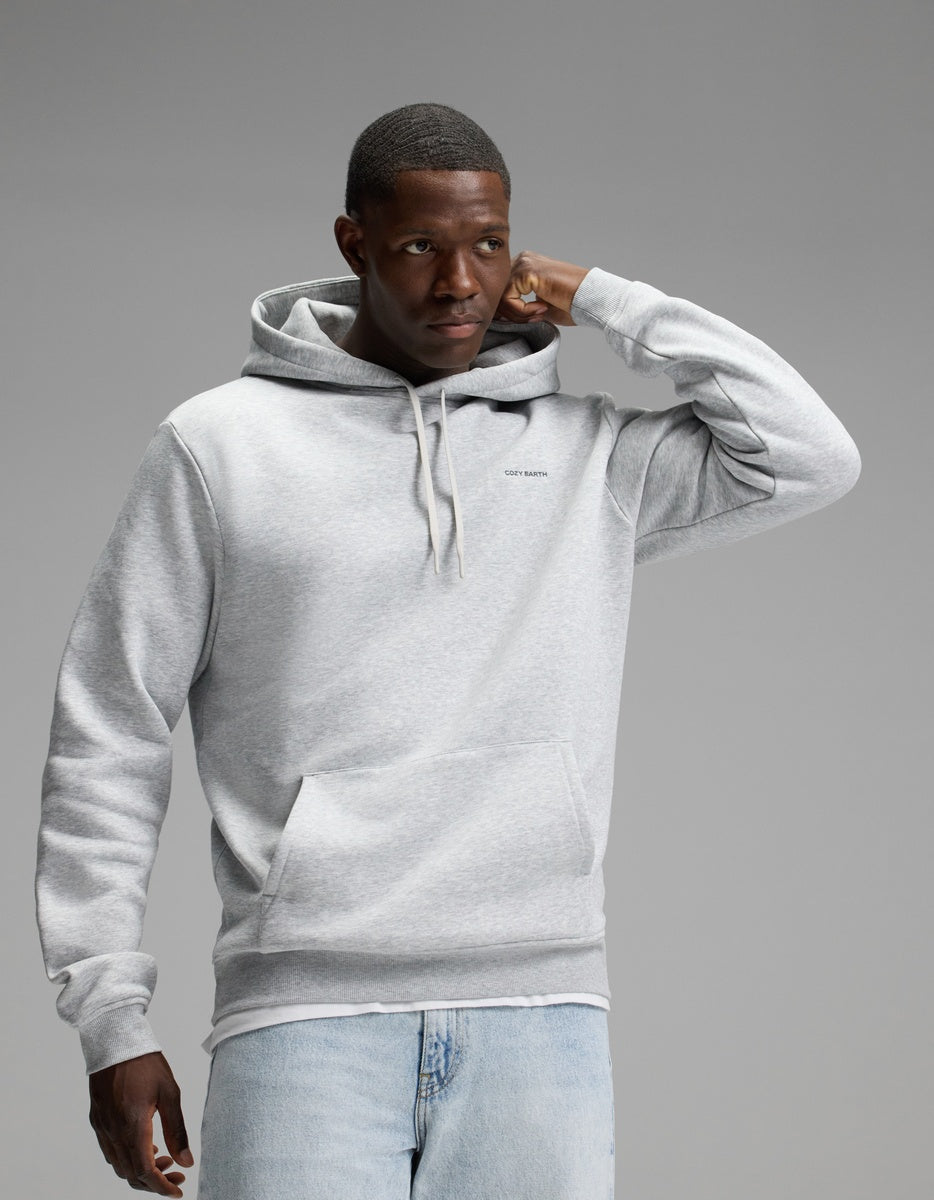 A man wearing the Cozy Earth Men’s Cityscape Hoodie in Heather Grey and light blue jeans stands against a plain gray background, with one hand touching his head and the other relaxed by his side.|Color:Heather Grey