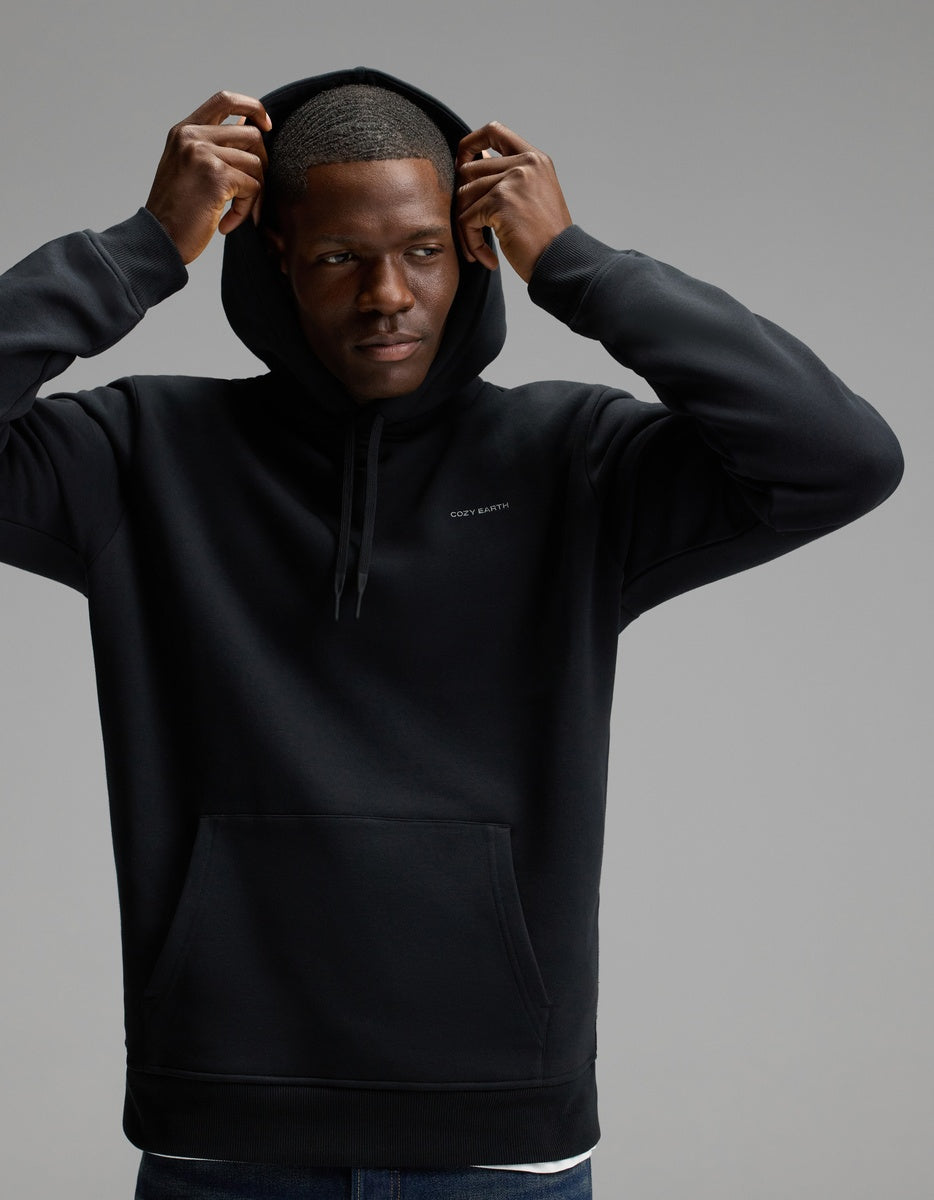 A man wears the Cozy Earth Men’s Cityscape Hoodie in Jet Black, lifting the hood over his head as he stands against a plain gray background, gazing to the side with a neutral expression.