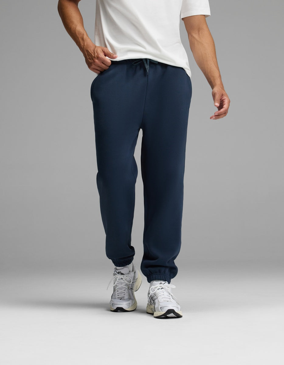 A person wearing a white T-shirt, white sneakers, and the Cozy Earth Men’s Cityscape Sweatpant in navy blue stands against a plain gray background. The image is cropped to show the individual from the shoulders down.