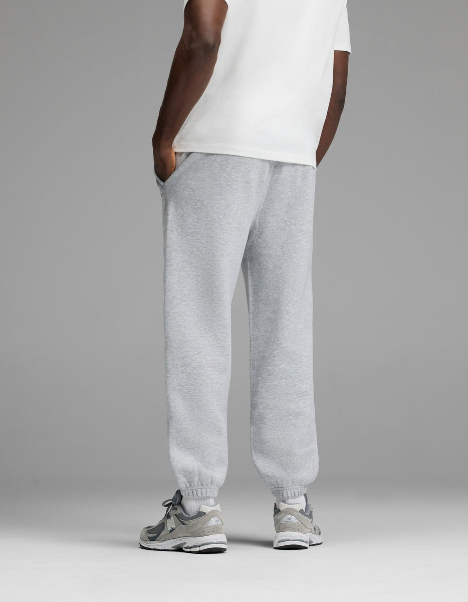A person stands with hands in pockets, wearing a white T-shirt, Cozy Earth Men’s Cityscape Sweatpant in light gray, and gray sneakers. The background is plain gray. The image is shown from behind the knees up.|Color:Heather Grey