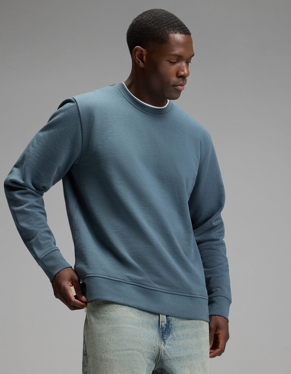 A man wearing the Cozy Earth Men’s Dylan Crewneck and light blue jeans stands against a plain grey background, looking down as he adjusts his sweatshirt.