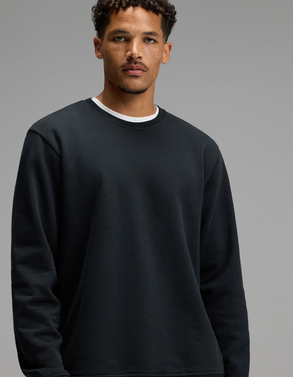 A man wearing the Men’s Dylan Crewneck in Jet Black by Cozy Earth over a white shirt stands against a plain gray background, looking toward the camera with a neutral expression.