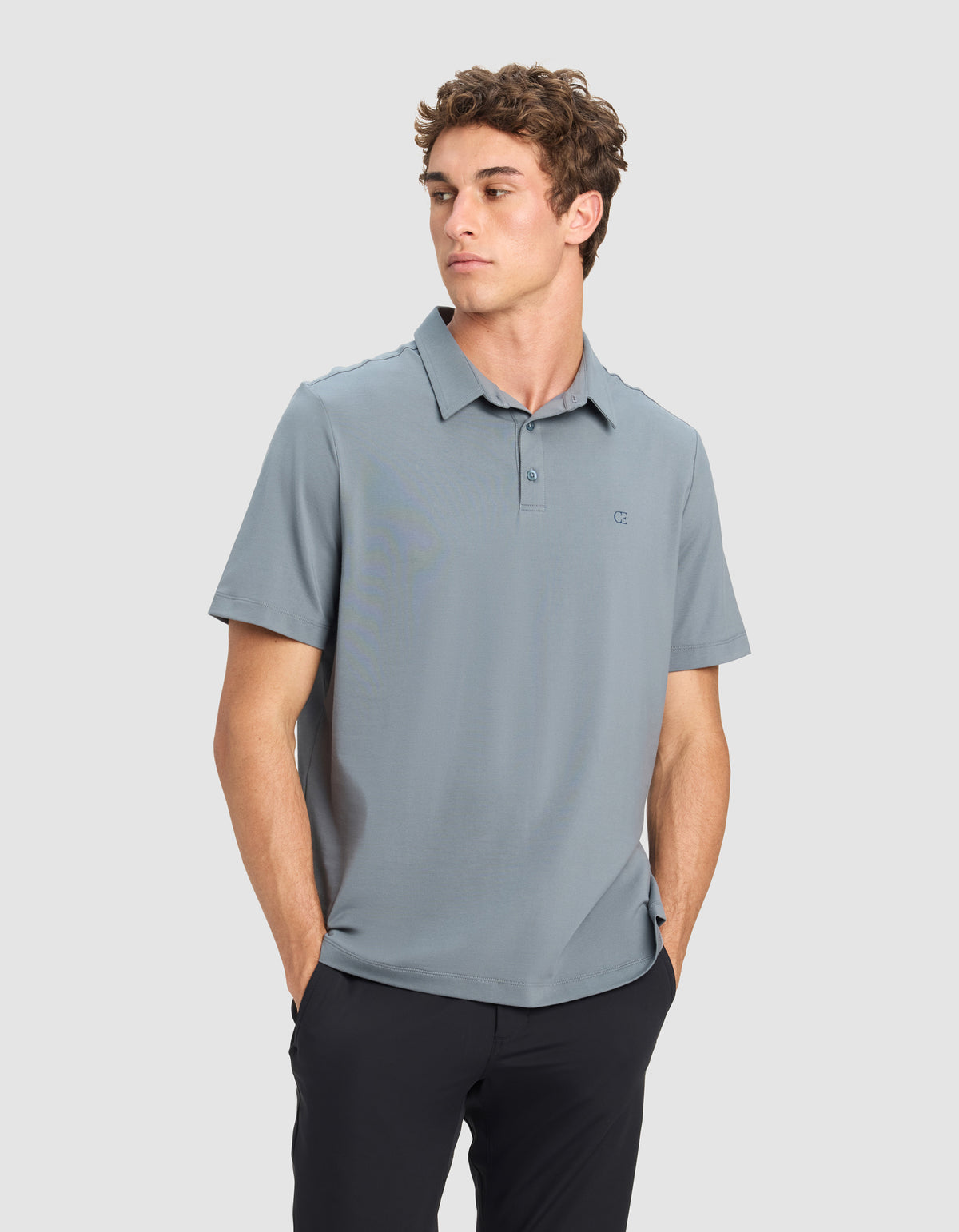 A young man wears the Cozy Earth Men's Everyday Polo in light blue and black pants, standing against a plain gray background with his hands in his pockets, looking to his left.