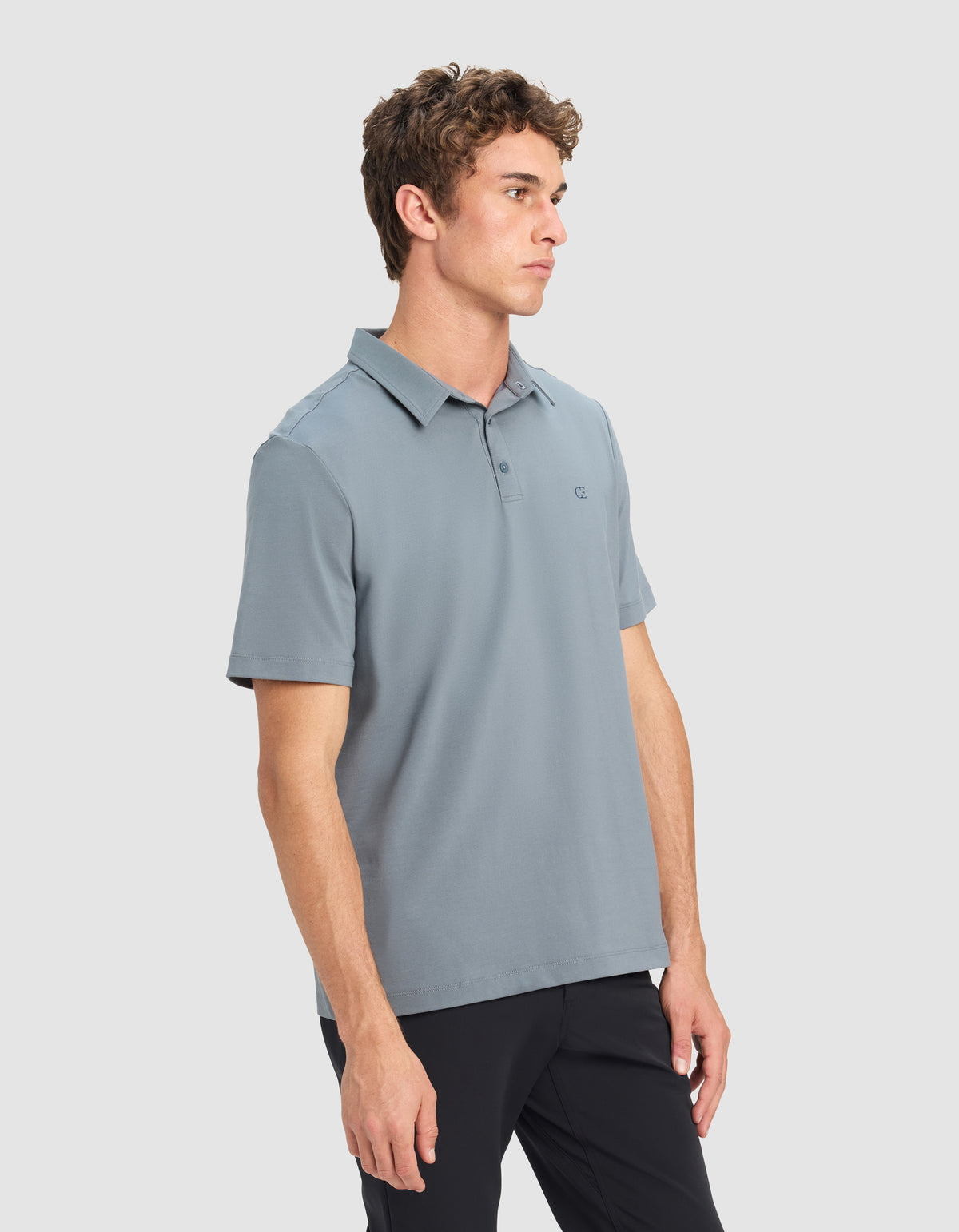 A young man with short curly hair models the Cozy Earth Men's Everyday Polo in light blue and black pants, standing against a plain light gray background and looking off to the side with a neutral expression.