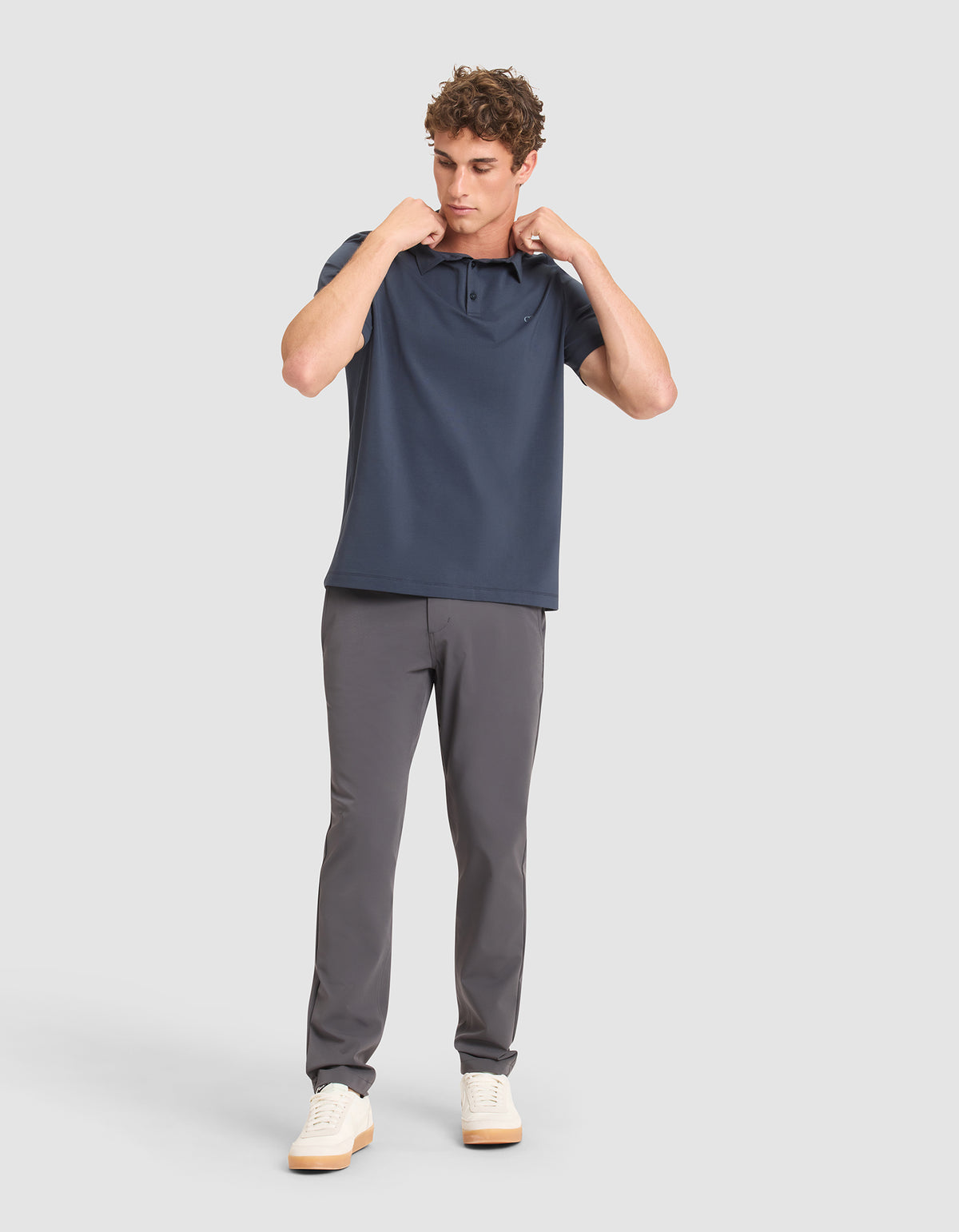 A man stands against a light gray background, wearing Cozy Earth's Men's Everyday Polo in navy with gray pants and white sneakers. He looks down while adjusting the collar of his shirt with both hands. 