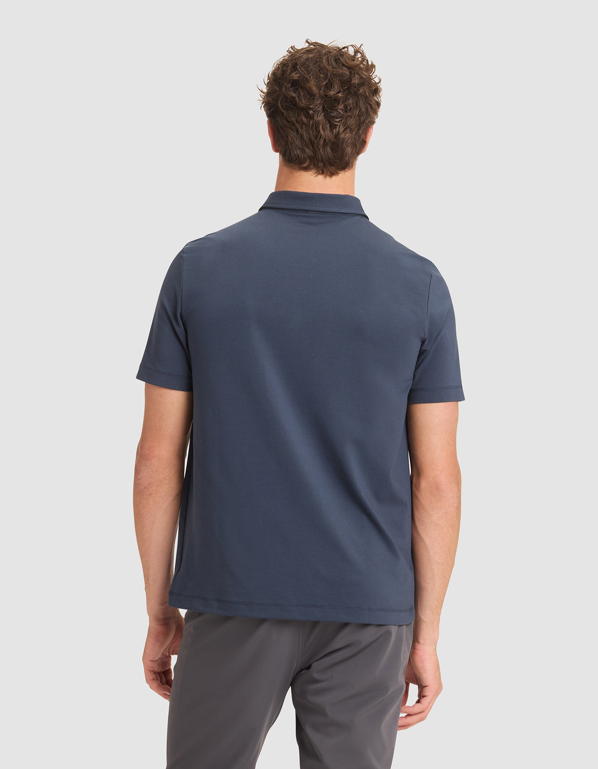 A man with short, curly brown hair is seen from behind wearing Cozy Earth's Men's Everyday Polo in navy blue and gray pants, standing against a plain light background. 