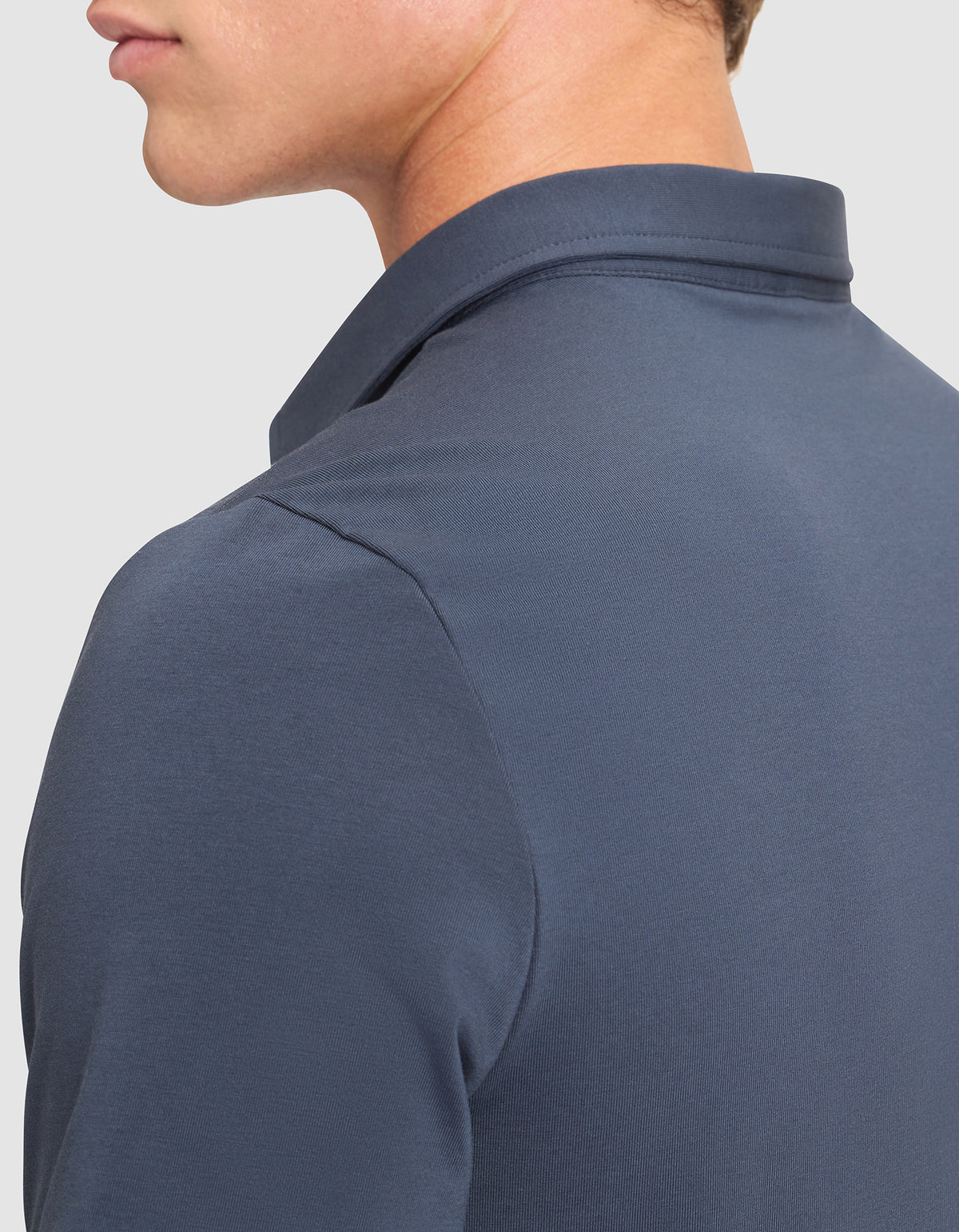 A close-up of a person wearing the Cozy Earth Men's Everyday Polo in dark blue, shown from the side and back. The image highlights the fabric, collar, and shoulder details with part of the head visible. 