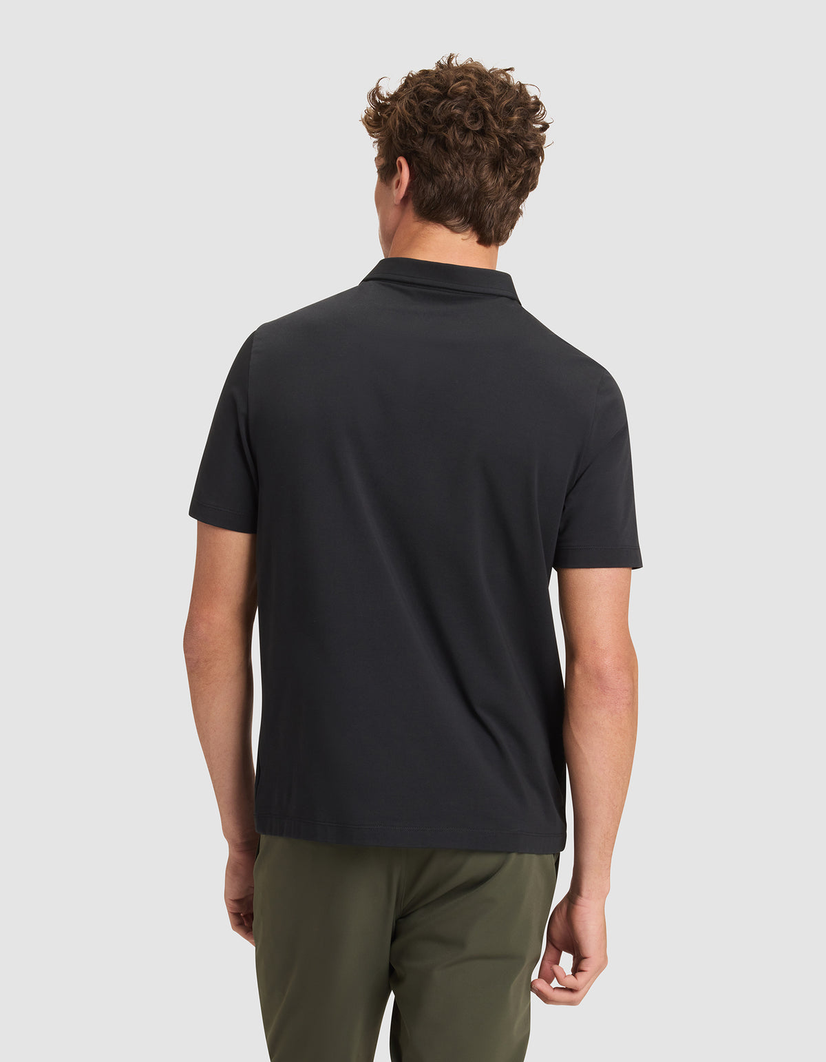 A man with curly brown hair faces away from the camera, wearing Cozy Earth's Men's Everyday Polo in black and olive green pants, set against a plain light gray background. 