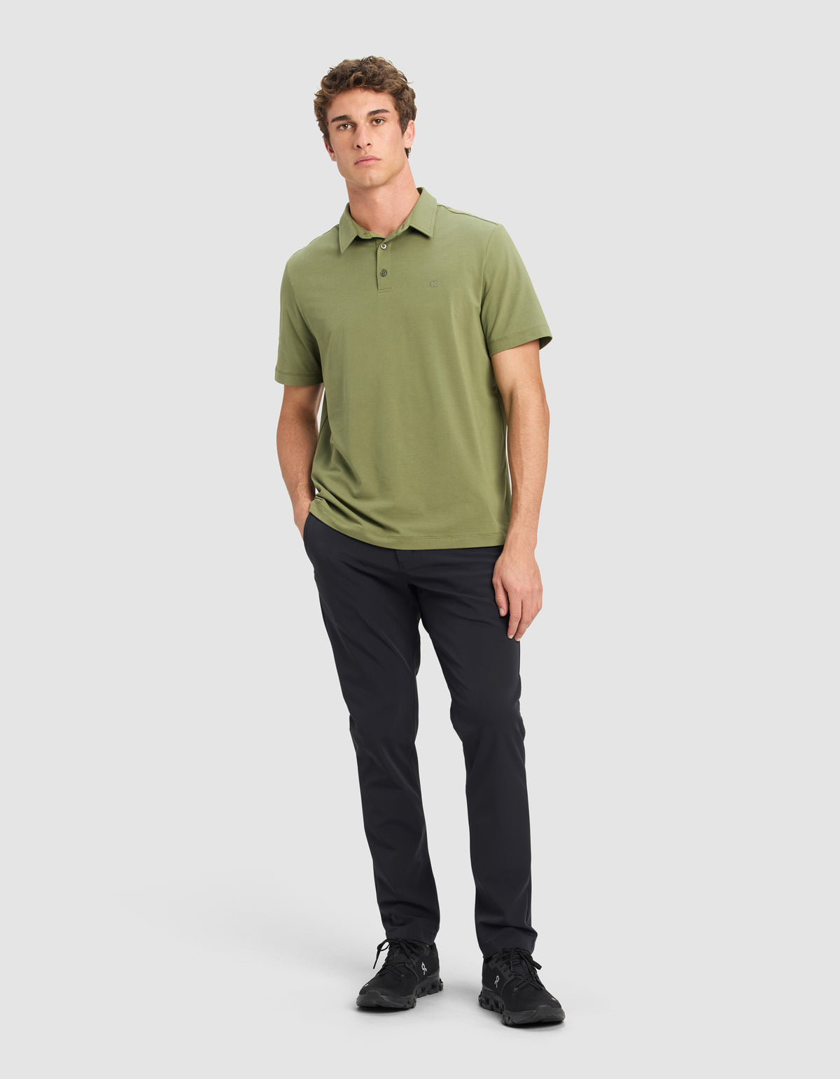 A young man faces forward with a neutral expression, left hand in his pocket, wearing the Cozy Earth Men's Everyday Polo in light olive green, paired with dark slim-fit pants and black sneakers against a plain background.