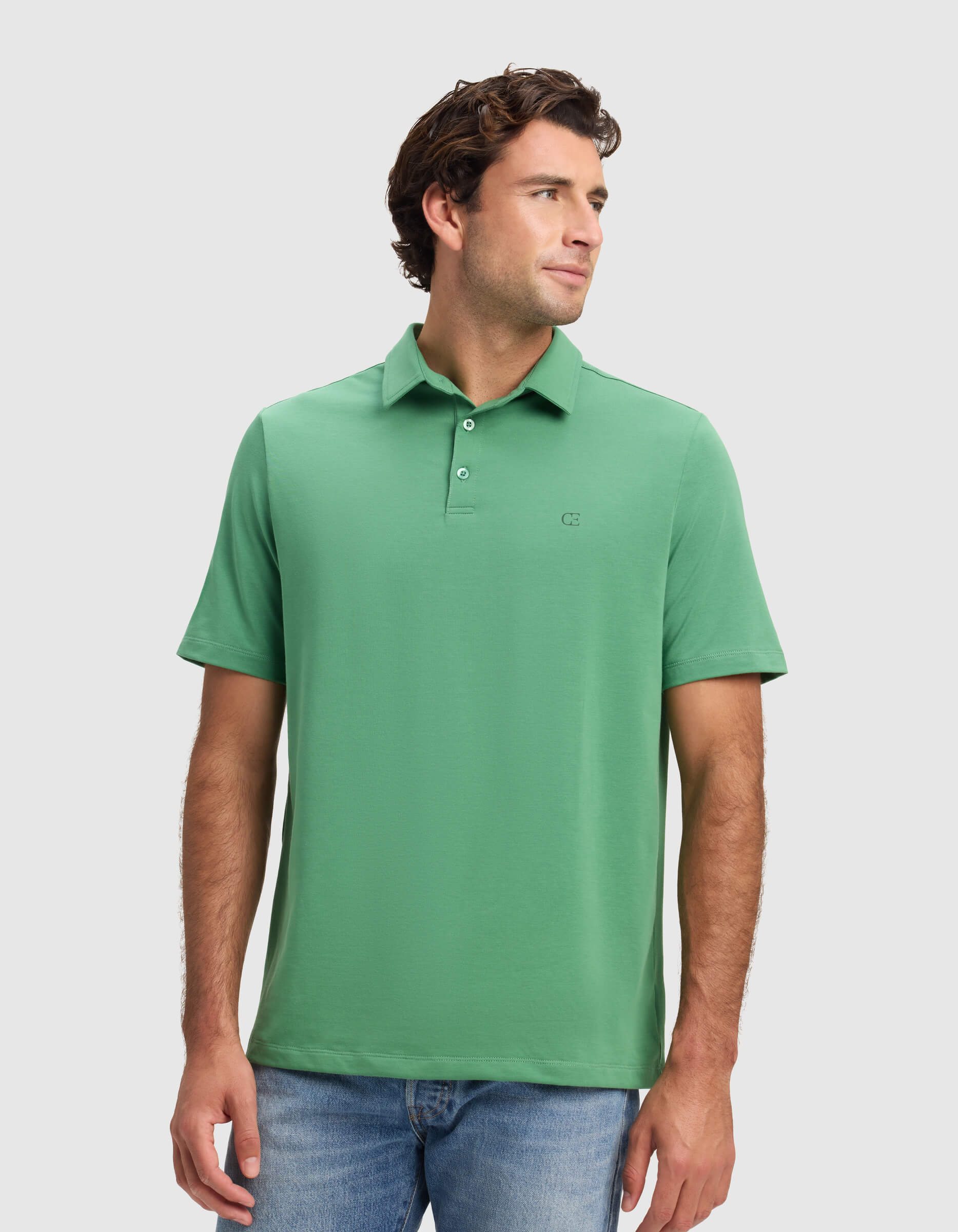 A man models the Cozy Earth Men's Everyday Polo in green with blue jeans, standing against a white background and gazing to his left with a relaxed expression.|Color:Vine