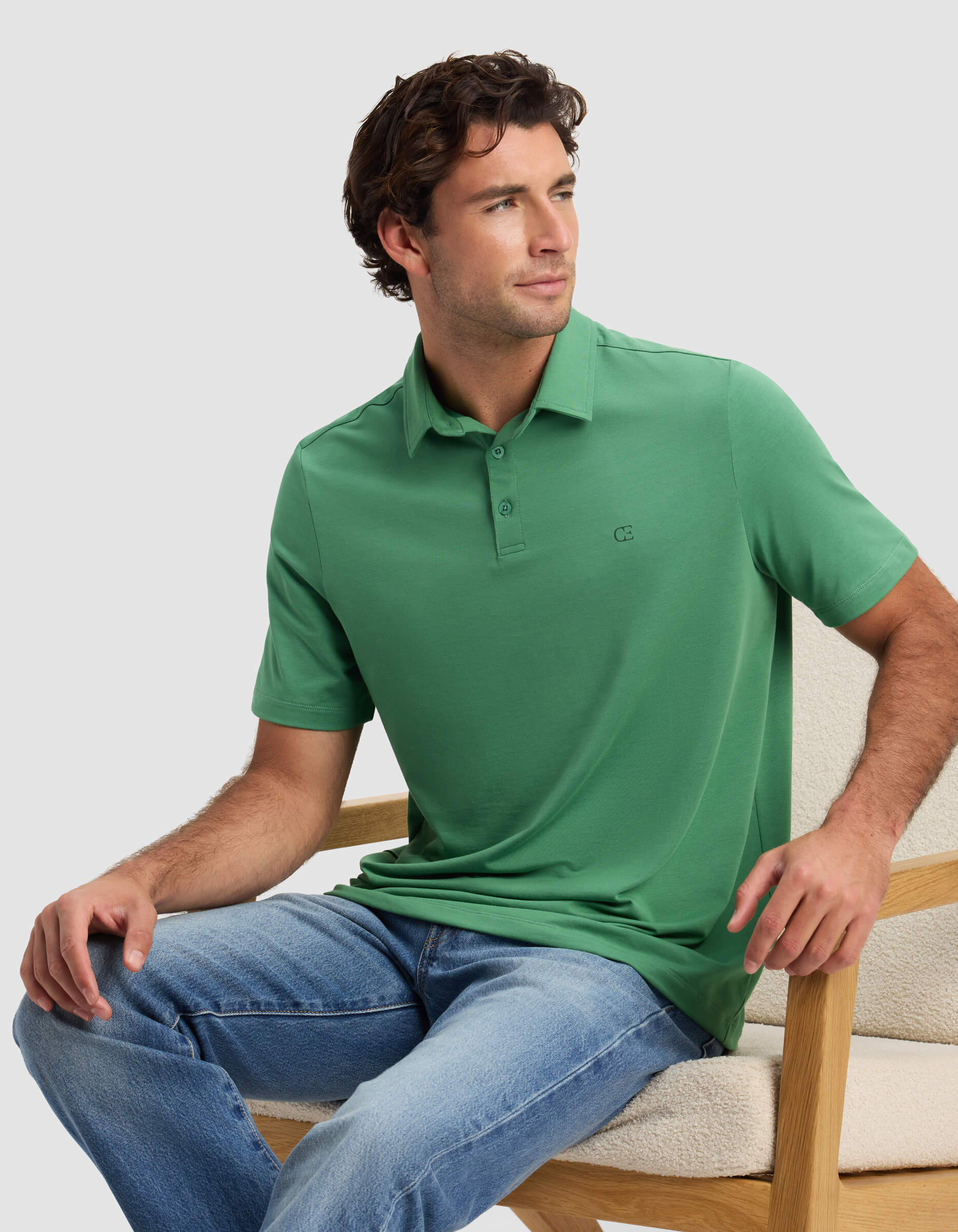 A man in a green Cozy Earth Men's Everyday Polo and blue jeans sits on a light wooden chair, gazing to the side with a relaxed expression.|Color:Vine