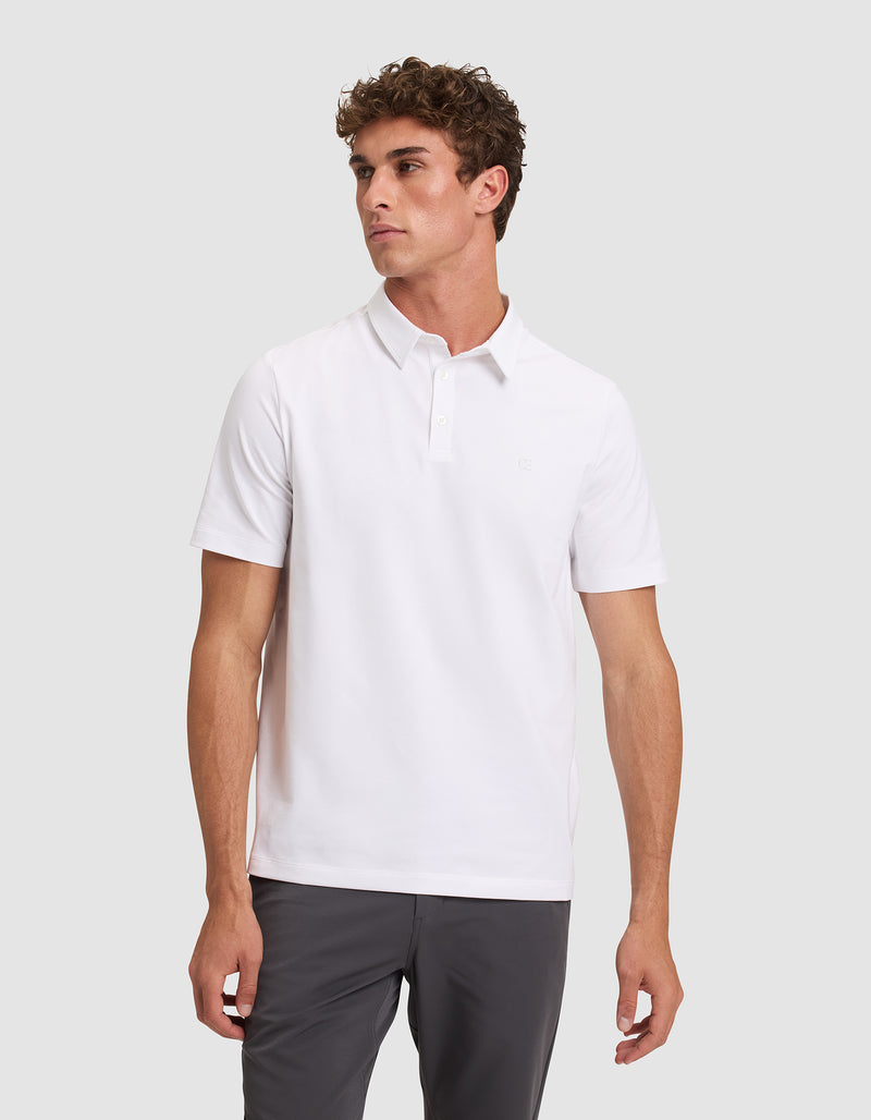A man with short curly hair wears the Cozy Earth Men's Everyday Polo in plain white and dark pants, standing against a light gray background and looking to his left with a neutral expression.