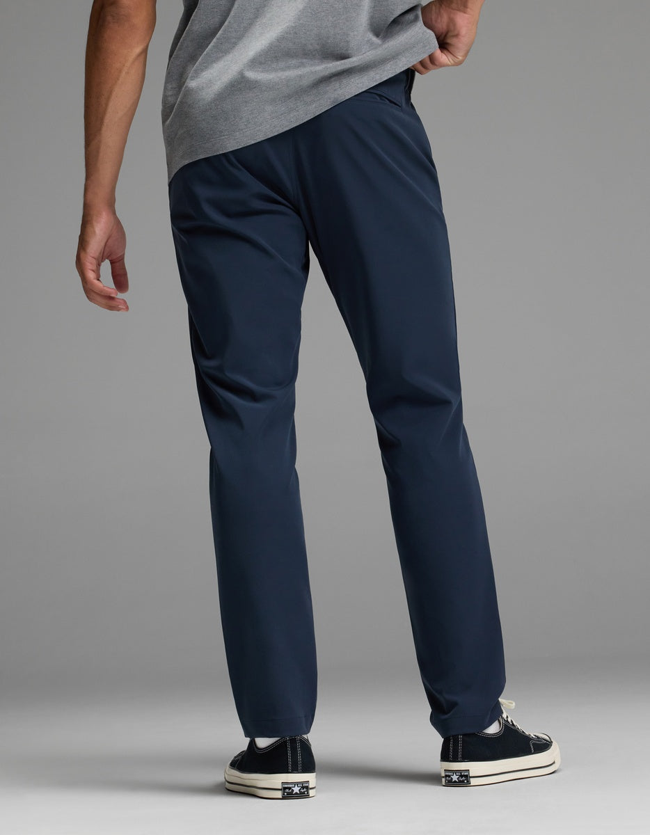 A man stands facing away against a plain background, wearing the Cozy Earth Men’s Everywhere Pant in navy blue, paired with a grey shirt and black sneakers with white soles, one hand resting on his hip.