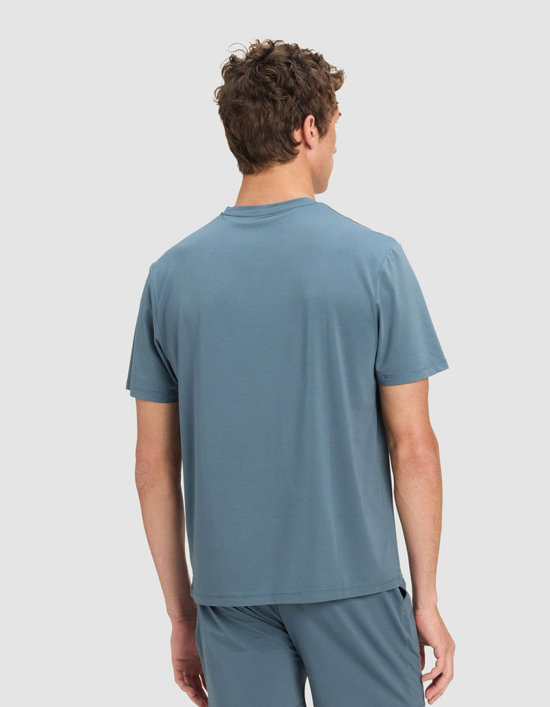 A man with short brown hair, seen from behind, is wearing the Cozy Earth Men's First Layer Pajama Short Sleeve Tee in light blue, paired with matching shorts against a plain light background.
