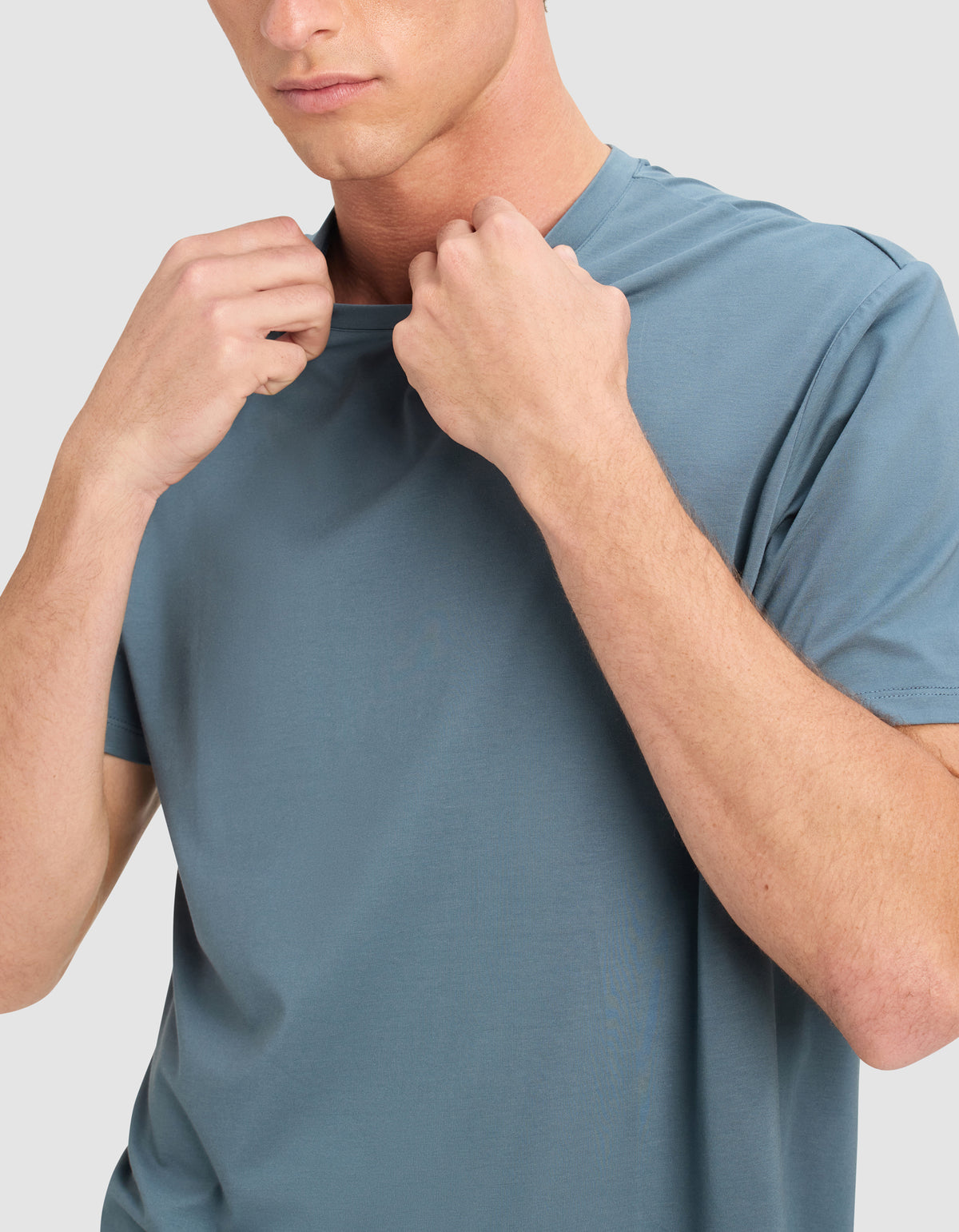 A person in a light blue Cozy Earth Men's First Layer Pajama Short Sleeve Tee holds the neckline with both hands. Their face is partially visible and neutral, set against a plain light background.