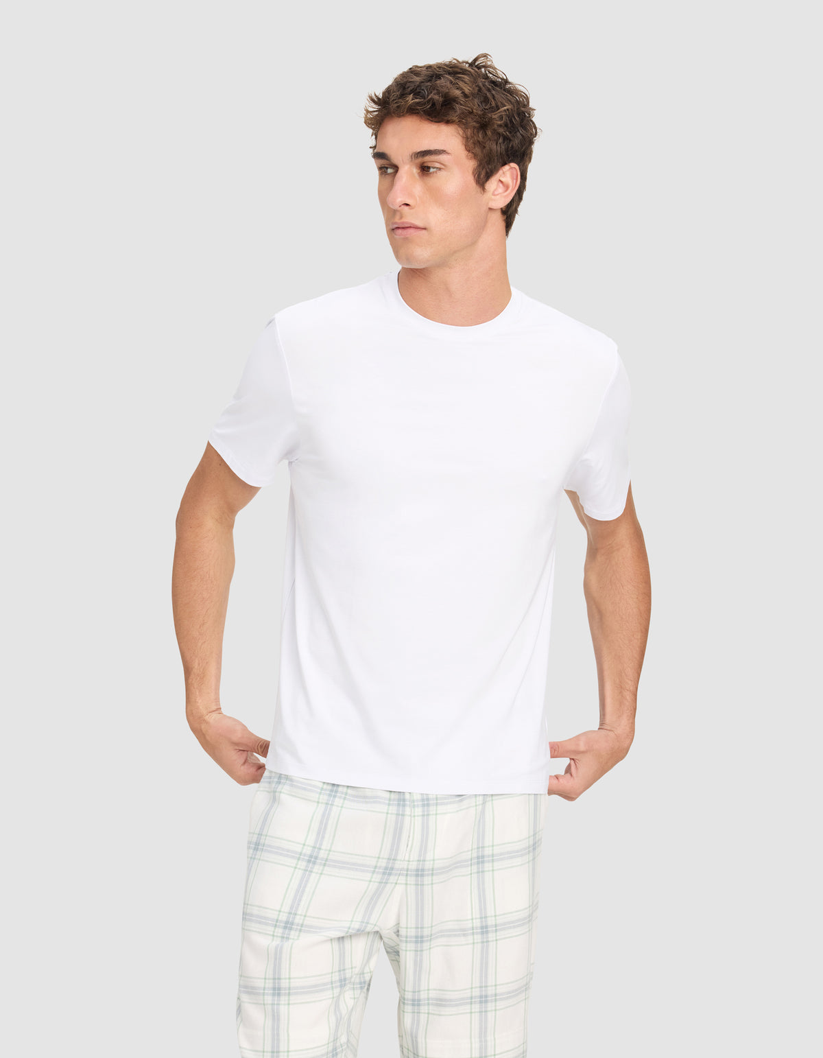 A young man models the Cozy Earth Men's First Layer Pajama Short Sleeve Tee and light plaid pajama pants against a light gray background, looking to his left while holding the hem of his shirt.