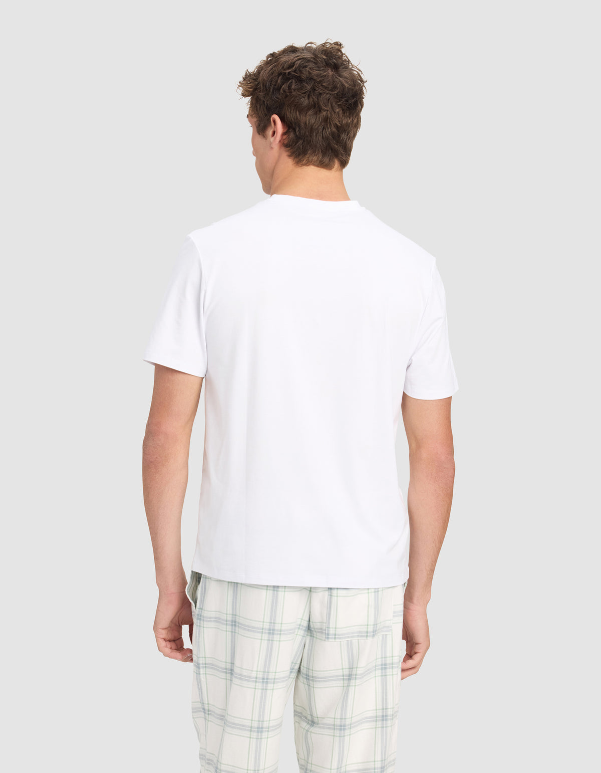 A man faces away from the camera, wearing the Cozy Earth Men's First Layer Pajama Short Sleeve Tee and light plaid shorts, set against a plain light gray background.