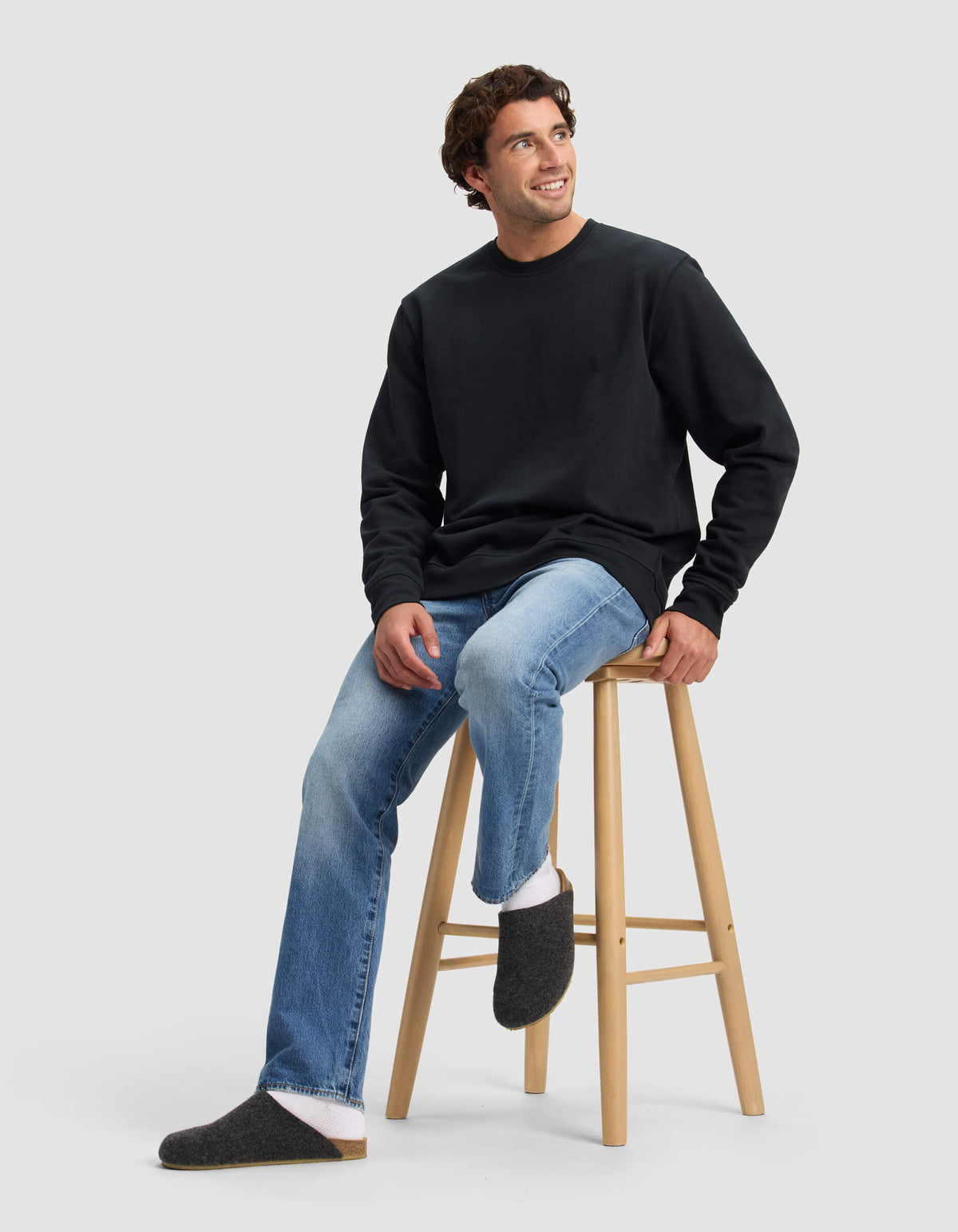 A man with curly brown hair sits on a wooden stool, smiling up, in a black sweatshirt, light blue jeans, white socks, and Cozy Earth Lakehouse Clog slippers. The backdrop is a plain light gray.|Color:Graphite