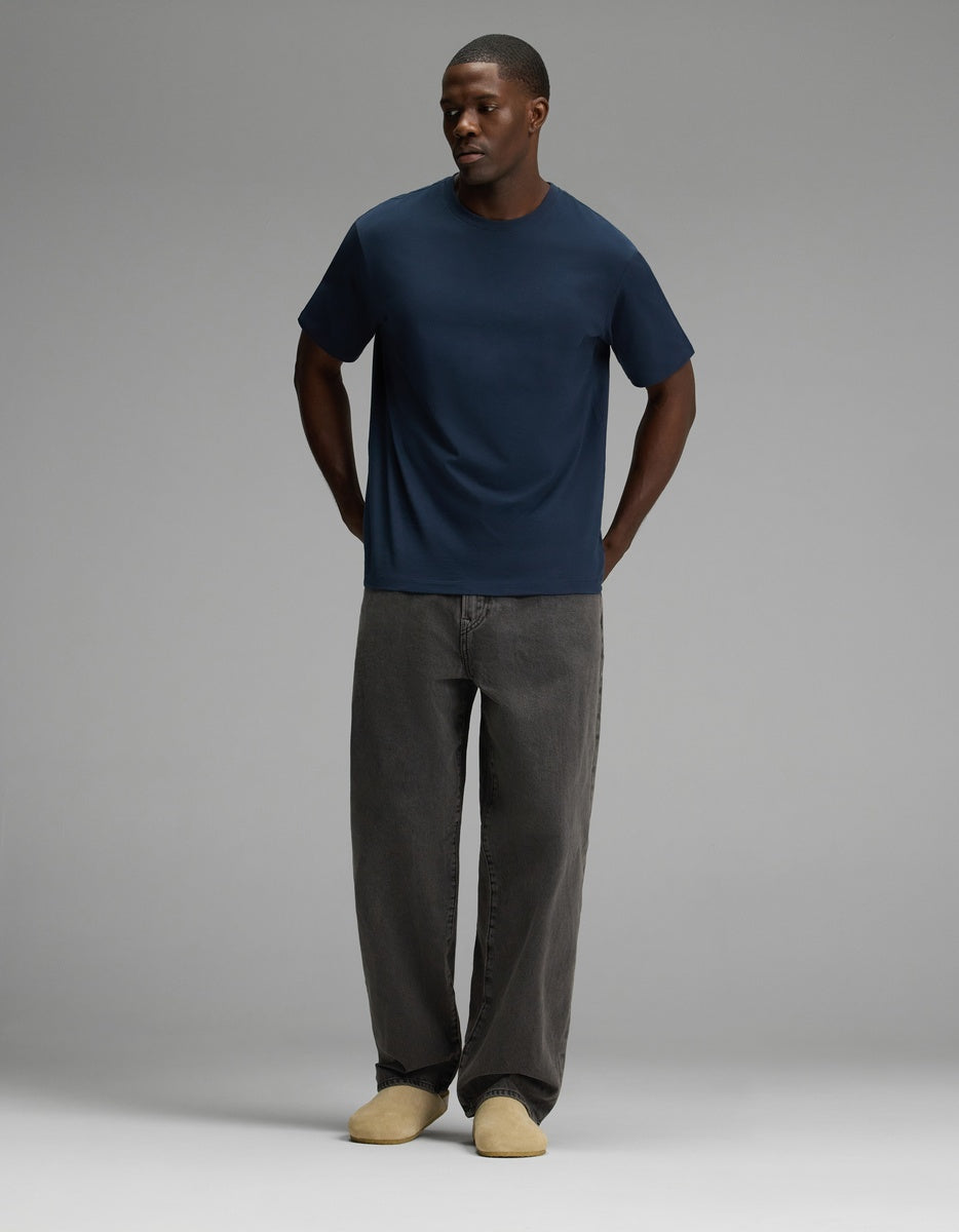 A person stands against a plain gray background, wearing a navy blue t-shirt, loose-fitting dark gray jeans, and the Lakehouse Clog in Suede by Cozy Earth, with their hands in their pockets.|Color:Sandalwood