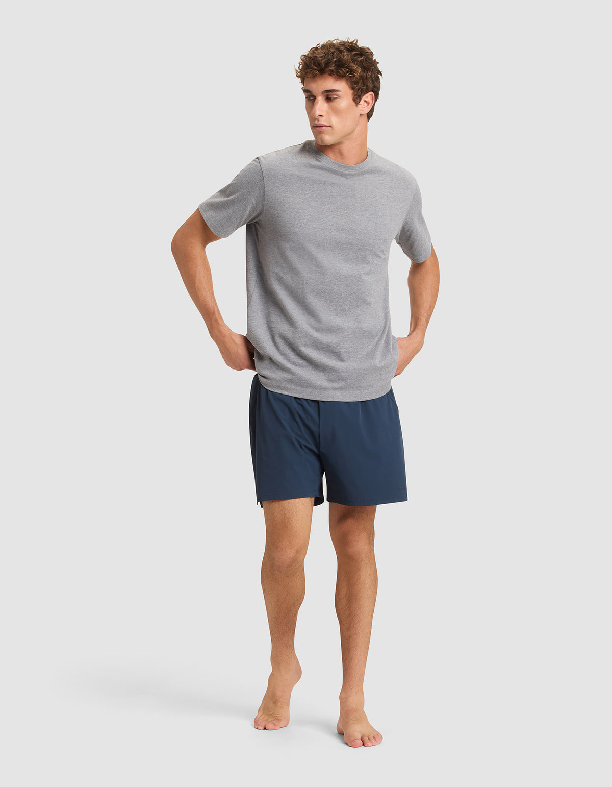 A man stands barefoot against a plain background, wearing a light gray short-sleeve T-shirt and Cozy Earth Men's Day & Night Shorts 5" in dark blue, with his hands resting on his hips. 