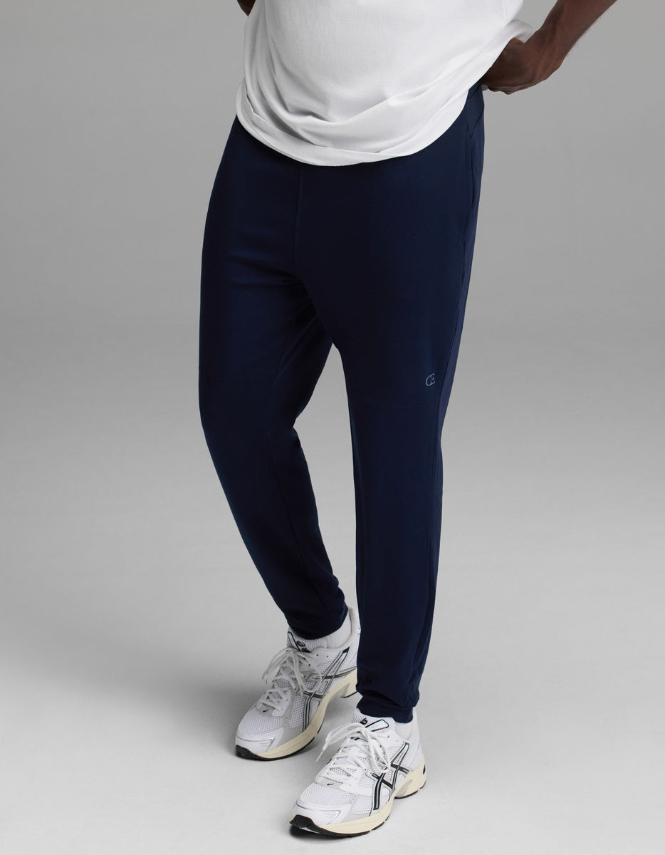 A person wearing a white shirt, Cozy Earth Men’s Recovery Jogger in navy, and white athletic sneakers stands against a plain gray background.