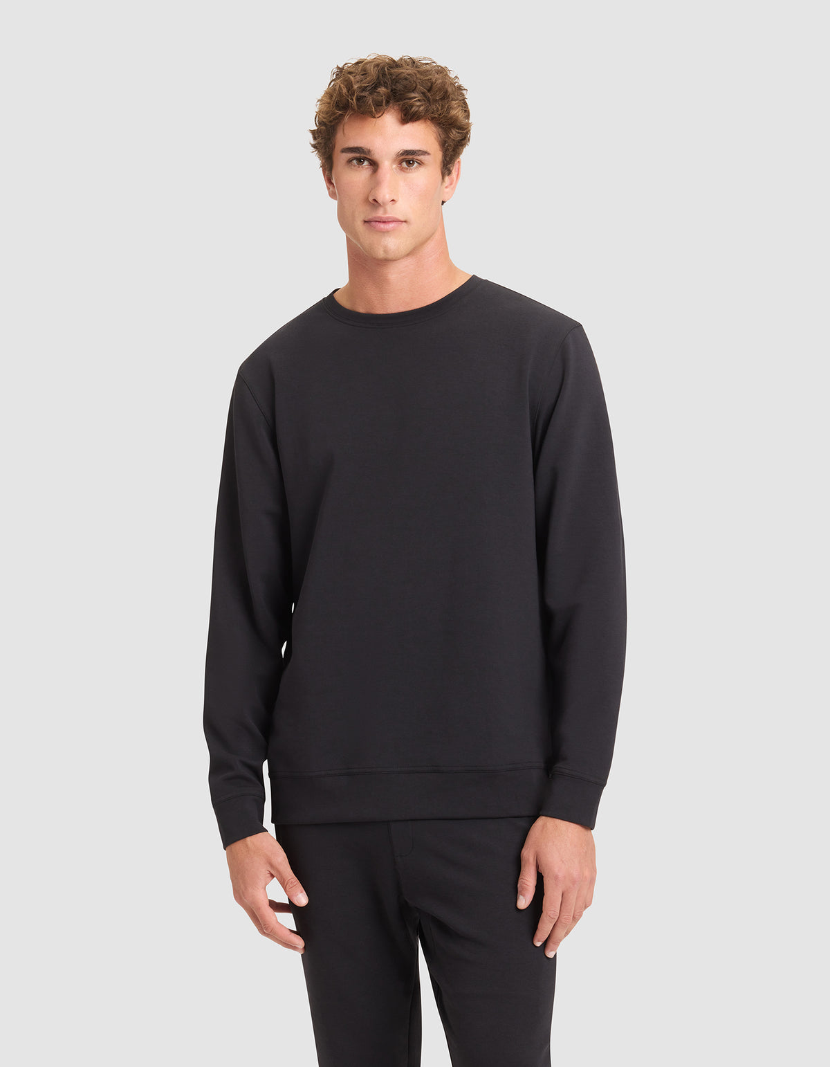 A young man with curly brown hair wears the Cozy Earth Men's StretchTech Crewneck in black with matching black pants, standing against a light gray background. 