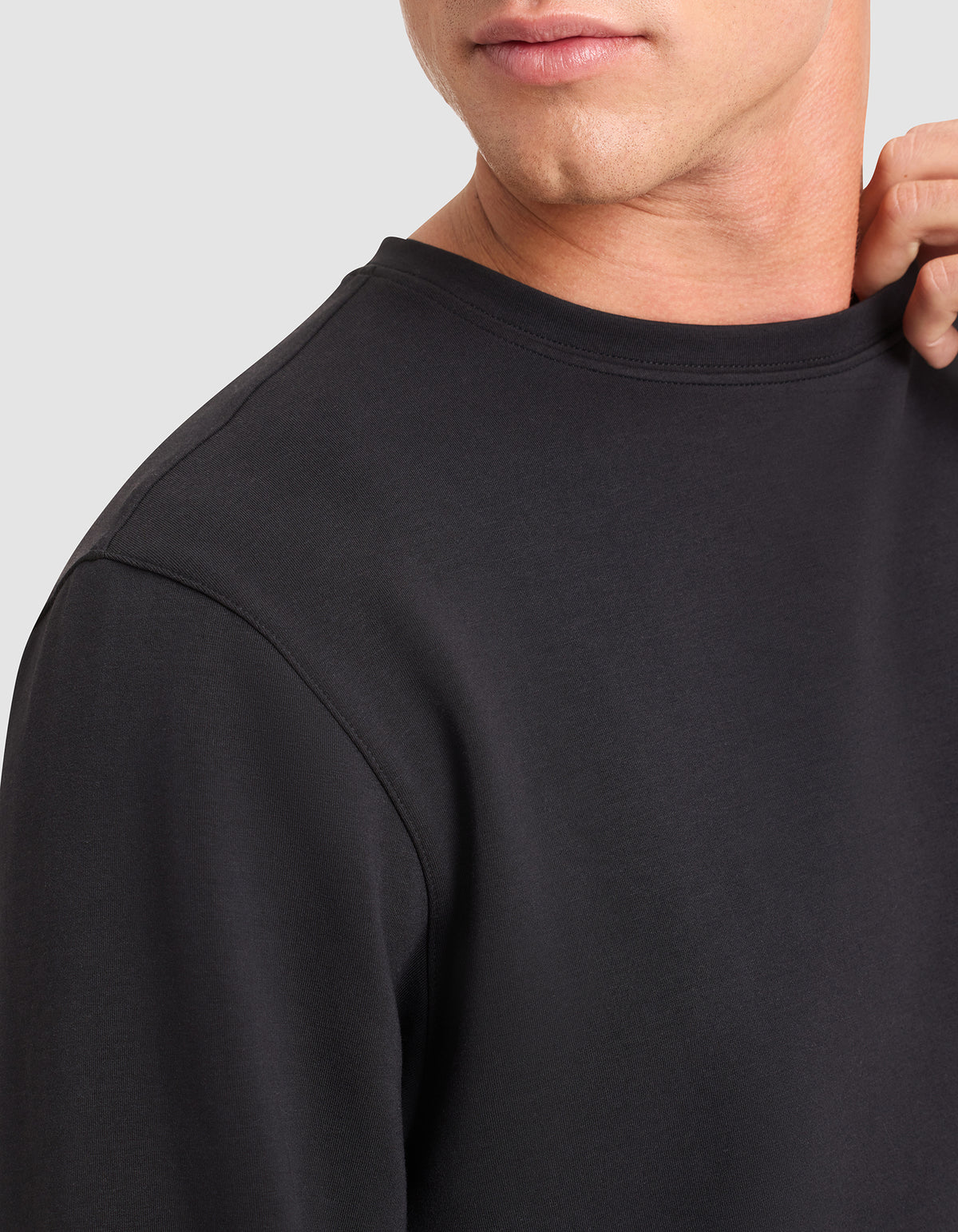 A close-up of a person wearing the Cozy Earth Men's StretchTech Crewneck in black, gently holding the collar. The image displays part of their face, neck, and upper torso. 