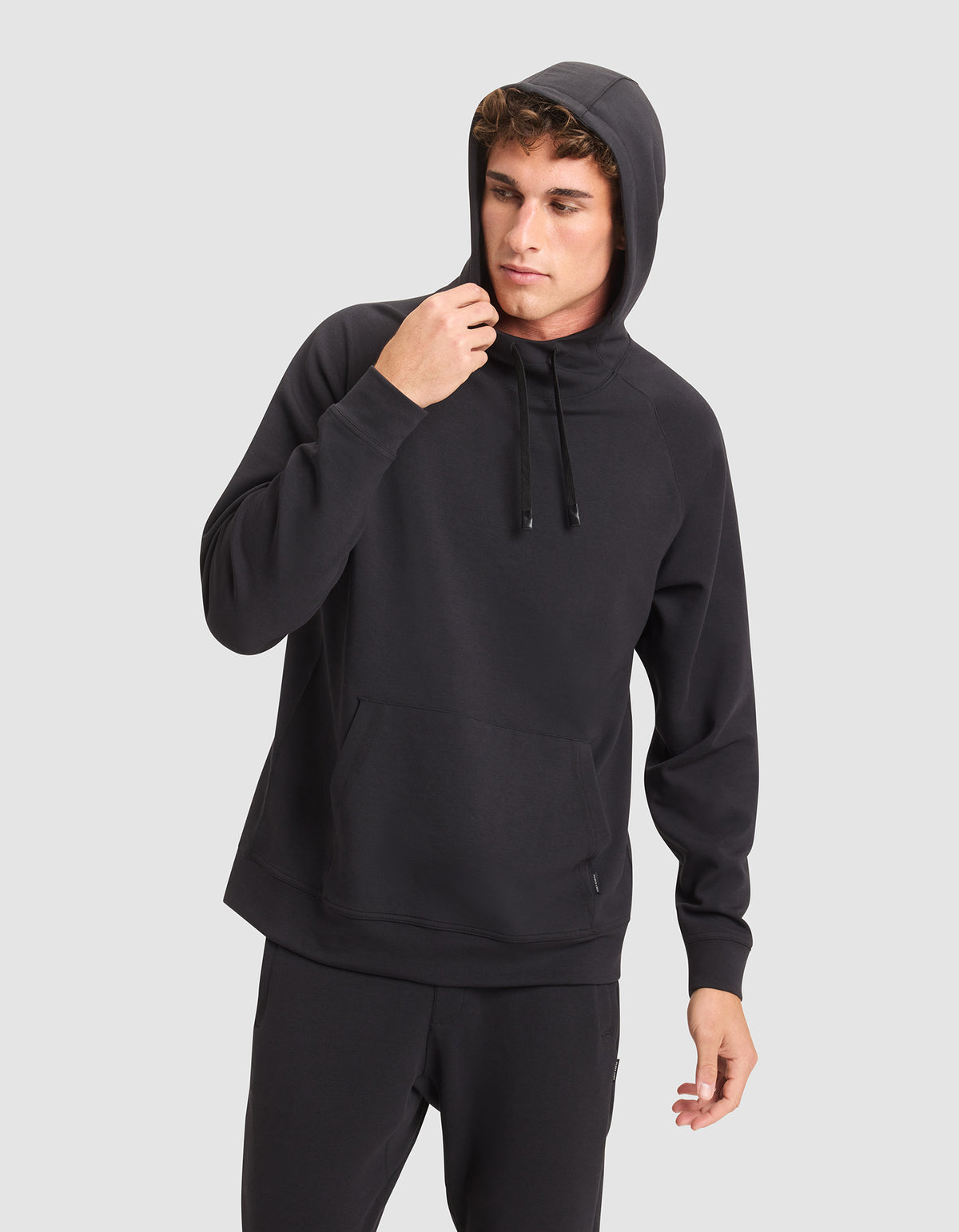 A young man wears the Cozy Earth Men's StretchTech Hoodie and black pants against a light gray background, looking to the side as he adjusts the hood. |Color:Jet Black
