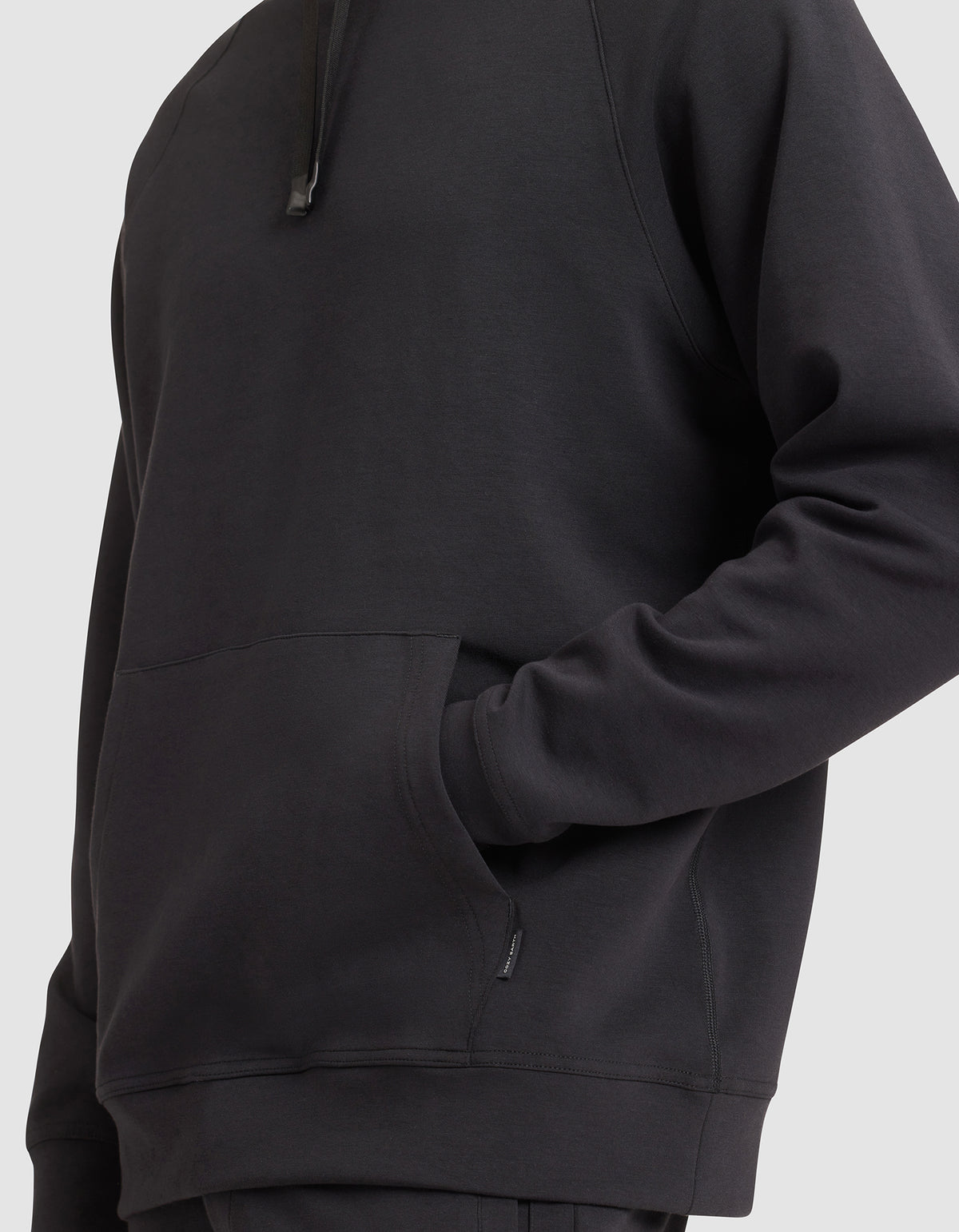 A man models the Cozy Earth Men's StretchTech Hoodie in black, one hand in the front pocket, shown from shoulders to hips against a plain light background. 