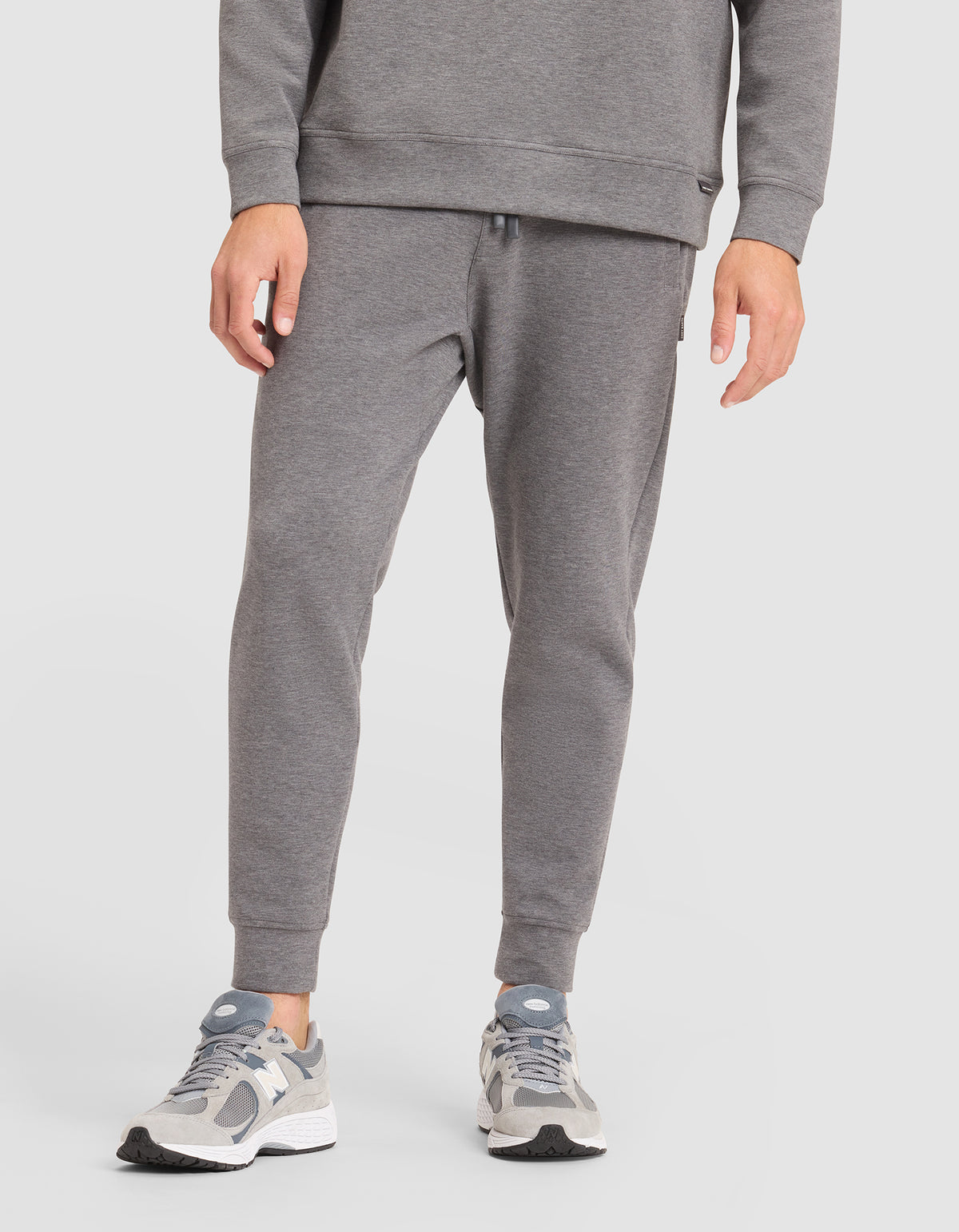 A person is shown from the waist down, wearing Cozy Earth's Men's StretchTech Jogger in gray with a matching sweatshirt and gray-white sneakers, standing against a plain white background. 