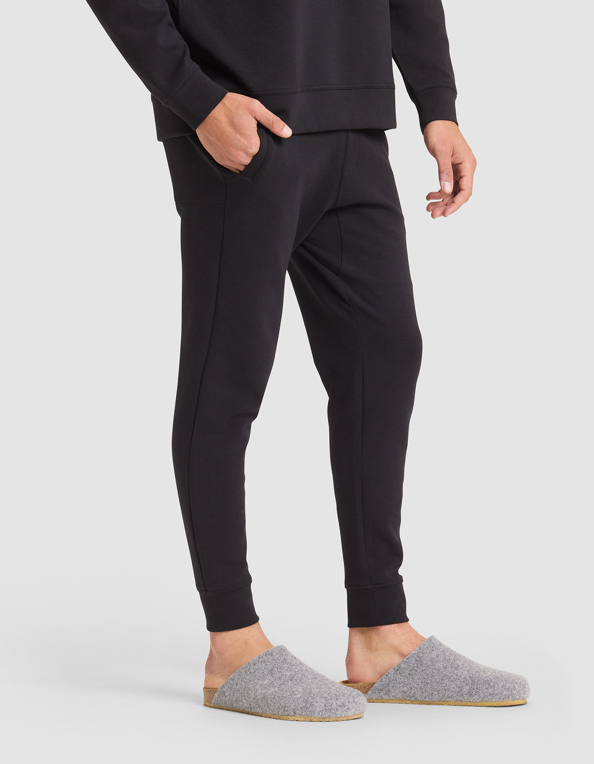 A person wearing Cozy Earth Men's StretchTech Jogger pants and a matching long-sleeve top stands with one hand in their pocket, wearing gray felt slippers with tan soles, on a white background. |Color:Jet Black