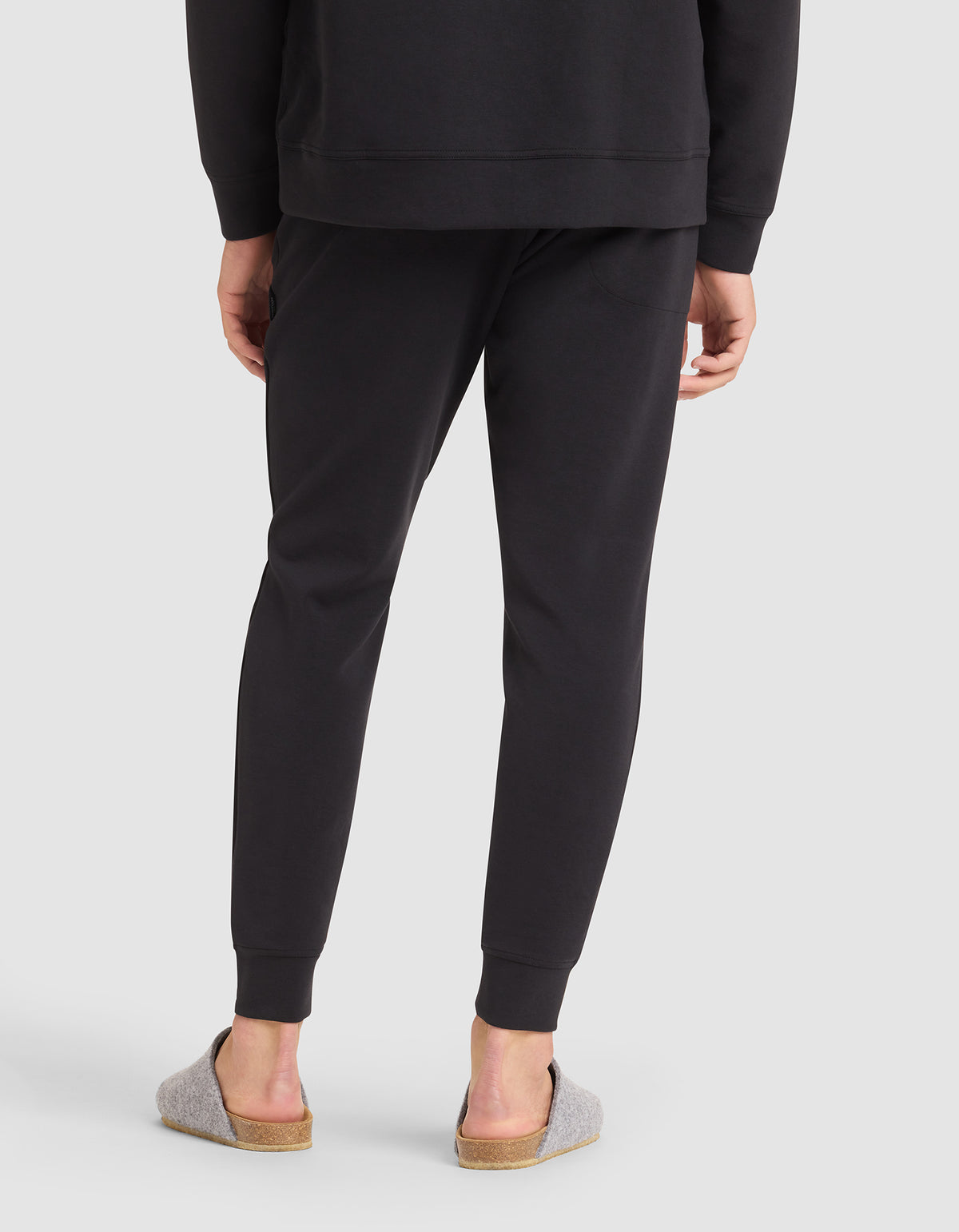 A person stands with their back to the camera, wearing Cozy Earth Men's StretchTech Jogger pants and a matching black sweatshirt, paired with light gray slippers with cork soles. The background is plain light gray. 