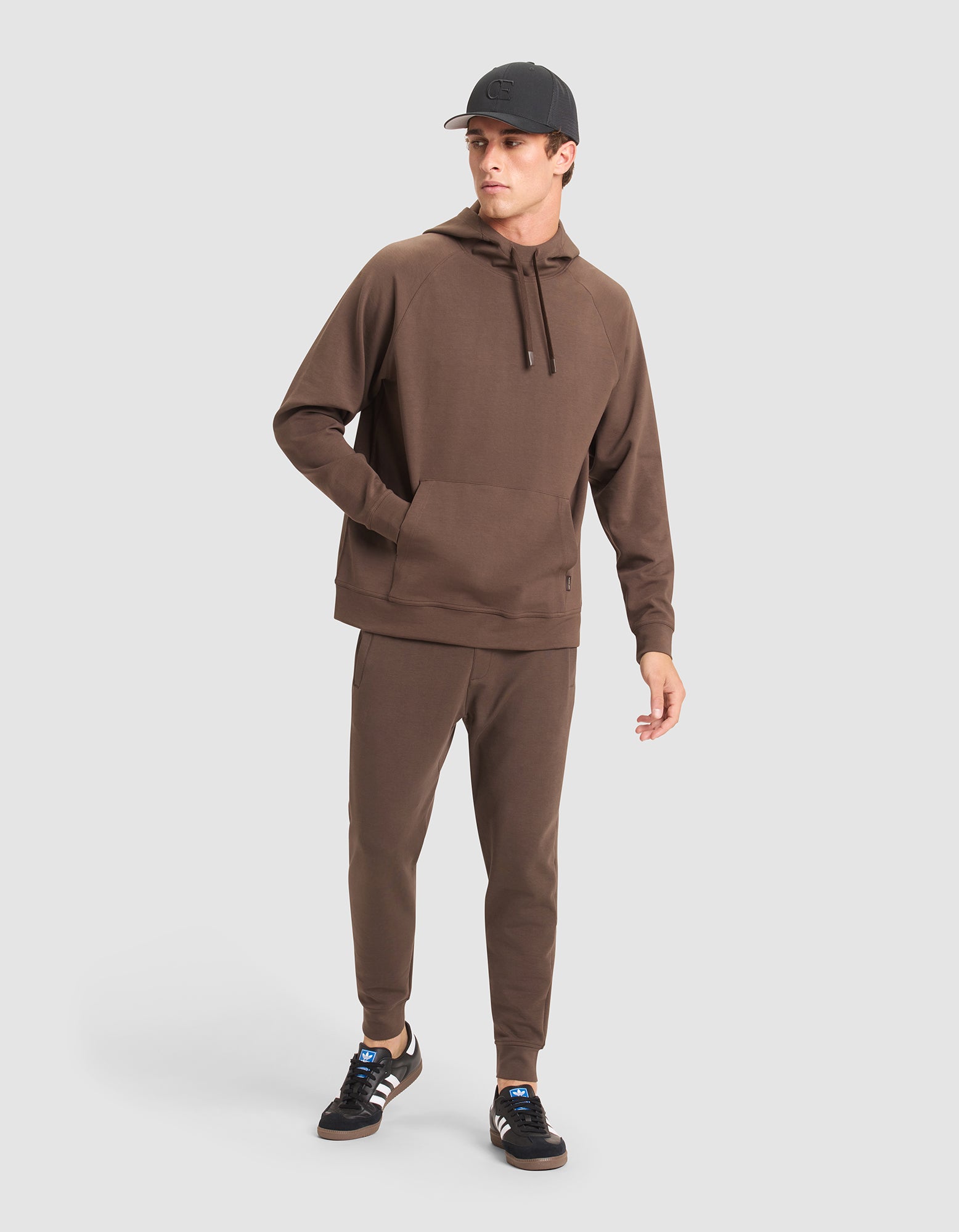A man wears the Cozy Earth Men's StretchTech Hoodie & Jogger Set with a black cap and black Adidas sneakers featuring white stripes, standing against a plain light gray background. |Color:Cacao