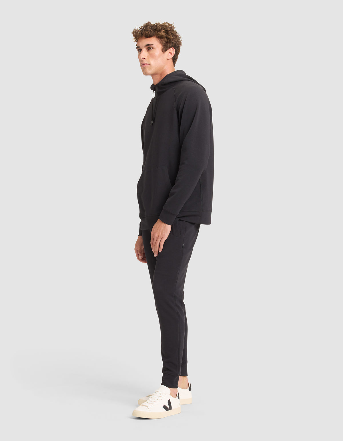A man stands sideways against a plain light gray background, modeling the Cozy Earth Men's StretchTech Hoodie & Jogger Set in black, paired with white sneakers featuring a black "V" logo. 
