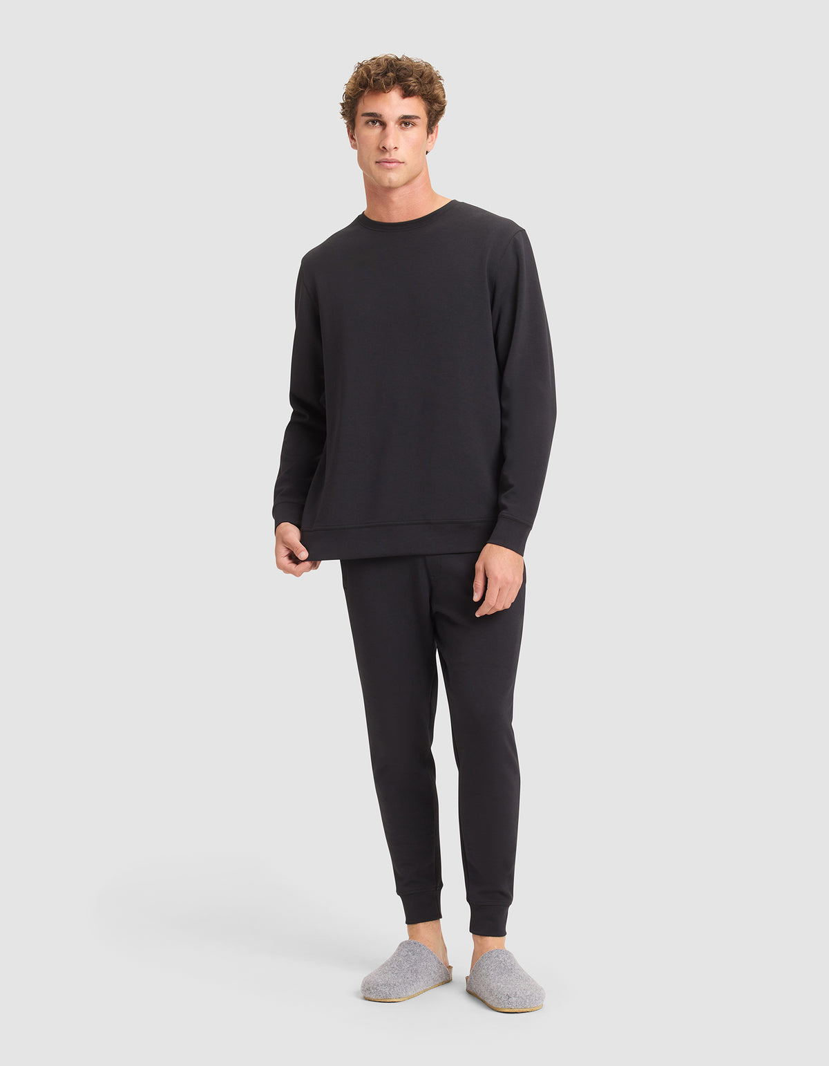 A man faces forward in a relaxed pose, wearing the Cozy Earth Men's StretchTech Crewneck & Jogger Set in black along with gray slippers, set against a plain background. |Color:Jet Black